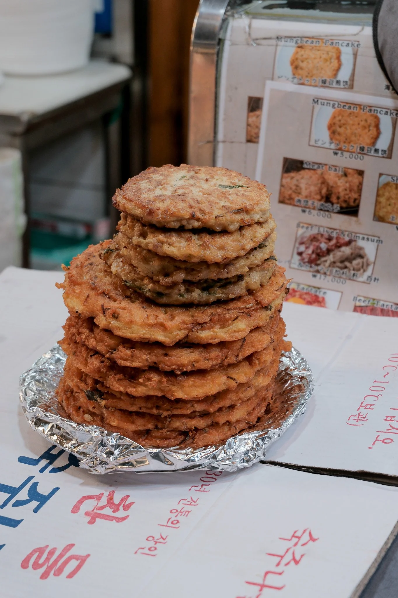 Ein Stapel knuspriger, goldbrauner koreanischer Mungbohnen-Pfannkuchen auf einer silbernen Alufolie, auf einem Tisch mit geschriebenen koreanischen Zeichen.