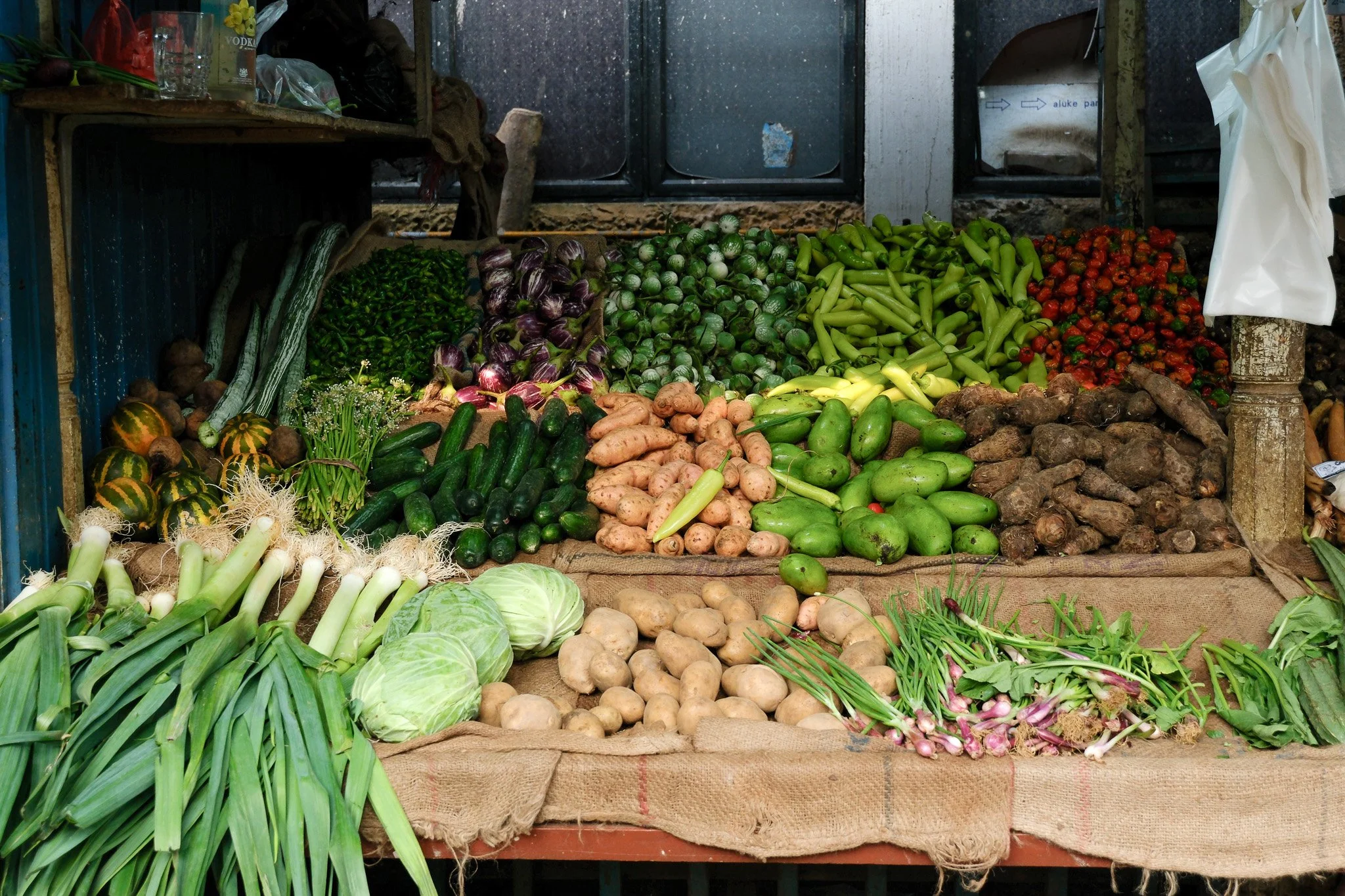 Verschiedene frische Gemüse auf einem Marktstand, darunter Zwiebeln, Kartoffeln, Salat, Zucchini, Gurken, Paprika, Tomaten, radieschen, und anderes Gemüse.
