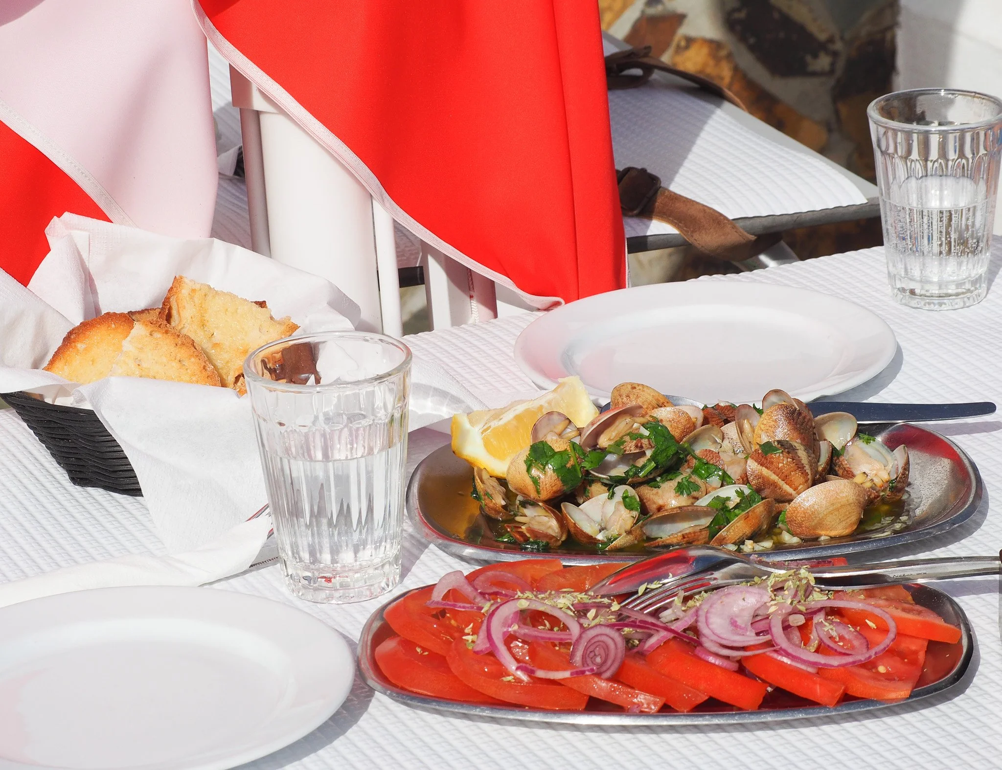 Ein gedeckter Tisch mit Meeresfrüchten, Tomaten, Zwiebeln und Zitronen, begleitet von Wasser und Brot, bei schönem Wetter