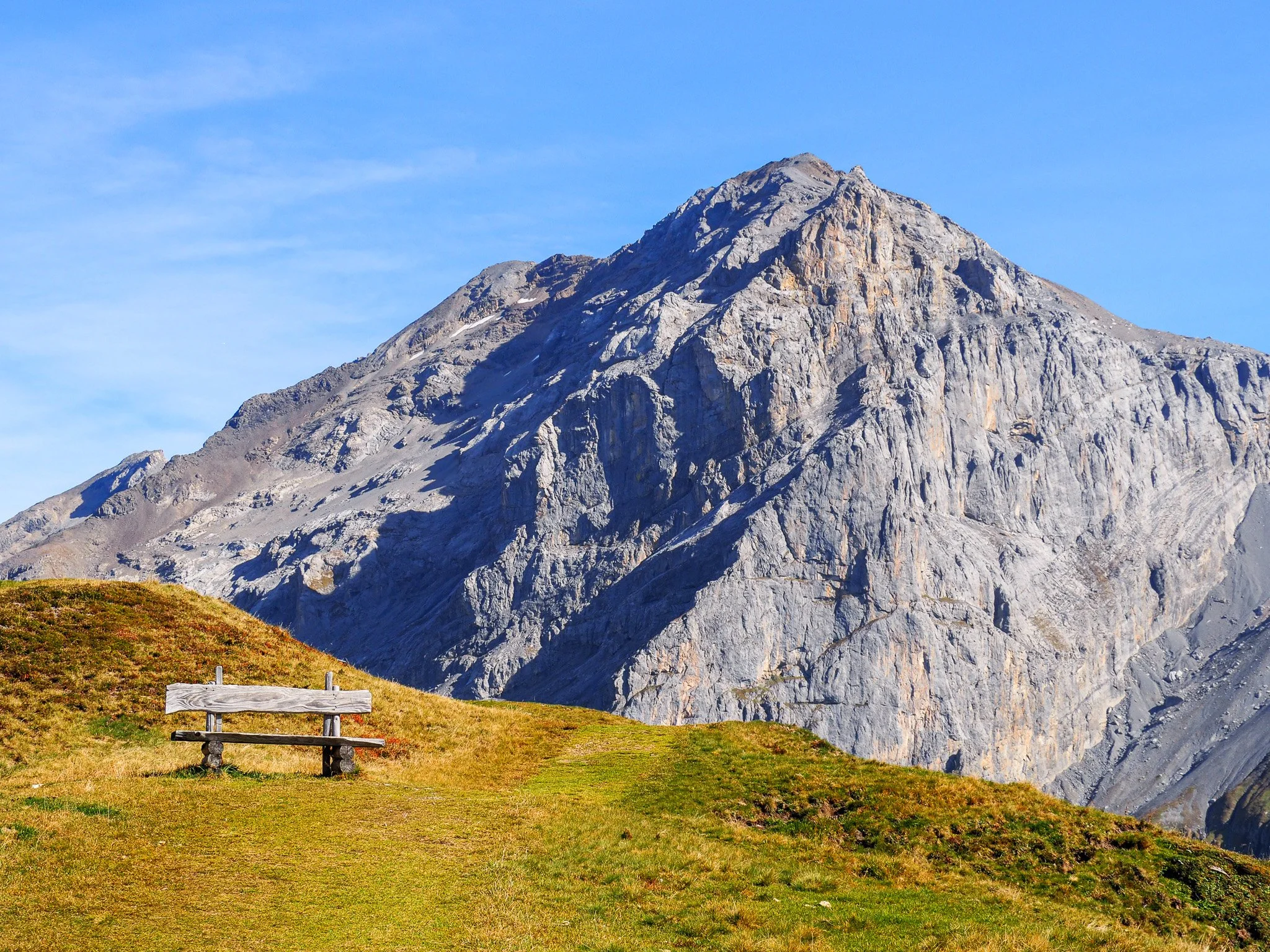 Ein einsamer Holzbalkon auf einer grasbewachsenen Wiese vor einer hohen, felsigen Bergwand unter einem blauen Himmel.