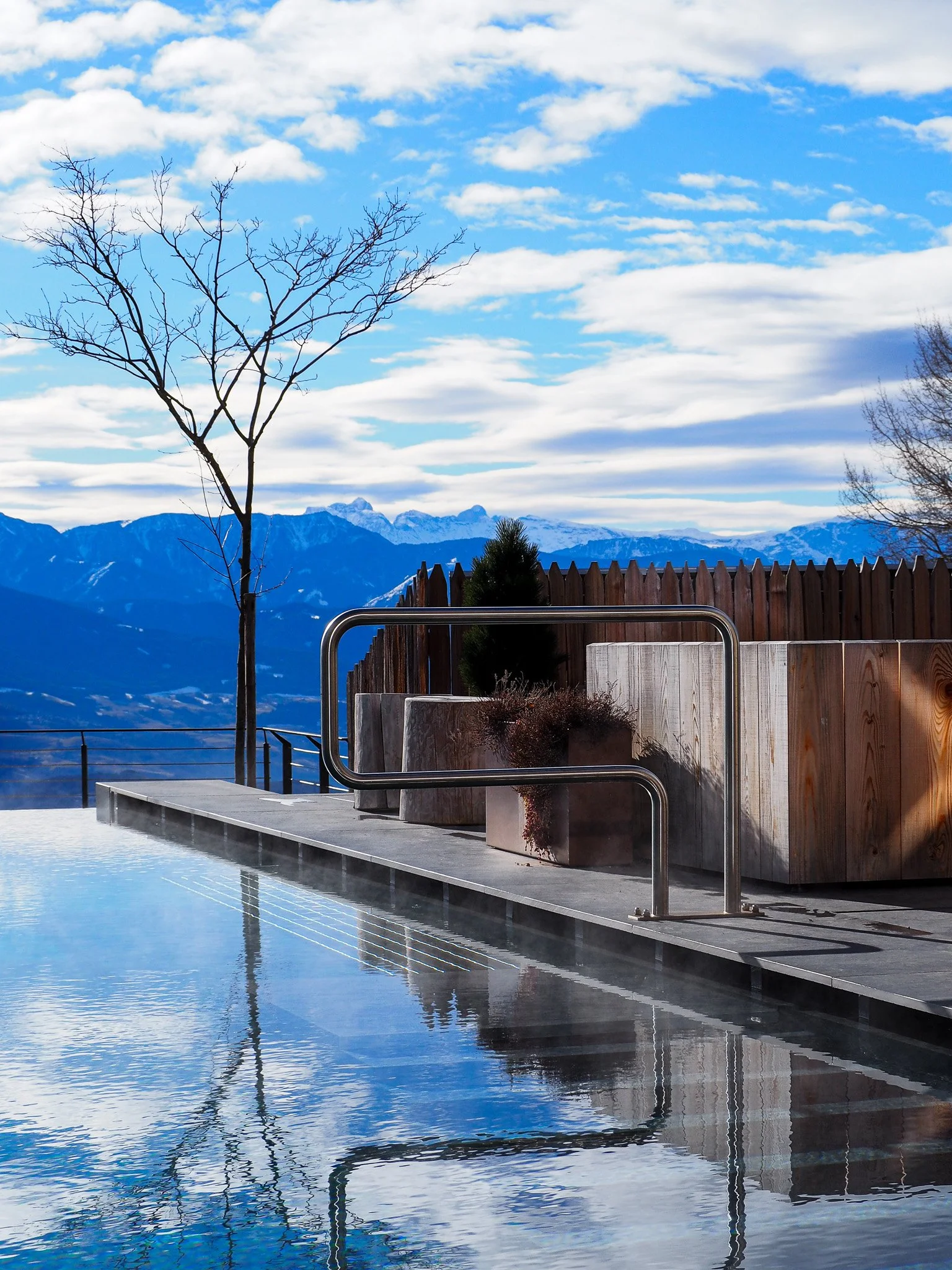 Berglandschaft mit Pool, Holzpflockzaun, Pflanzen in Behältern, nacktem Baum, und schneebedeckten Bergen im Hintergrund unter einem bewölkten Himmel.