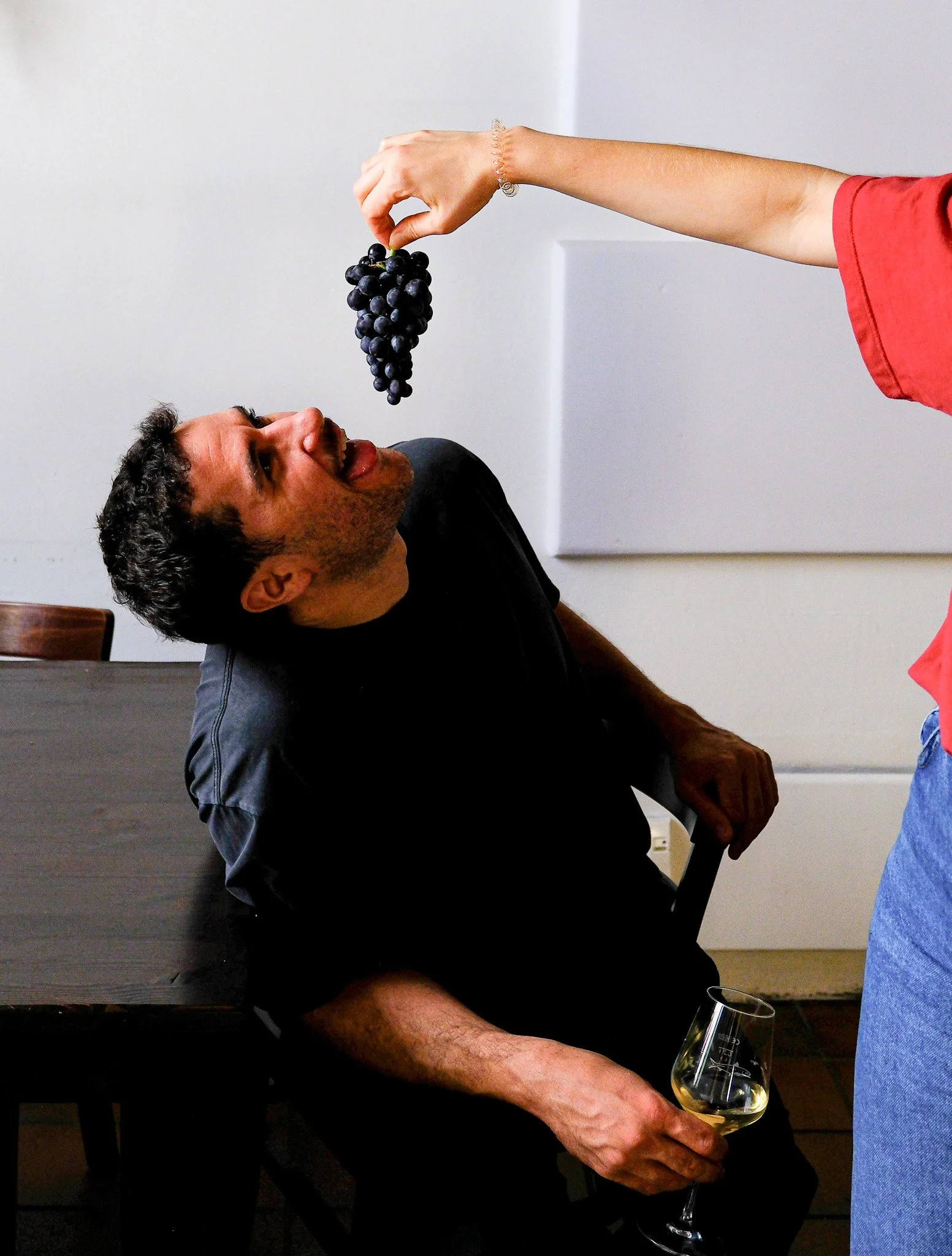 Jemand hält eine Traube Wein über den Mund eines Mannes, der in einem Raum sitzt, während er ein Weißweinglas hält.