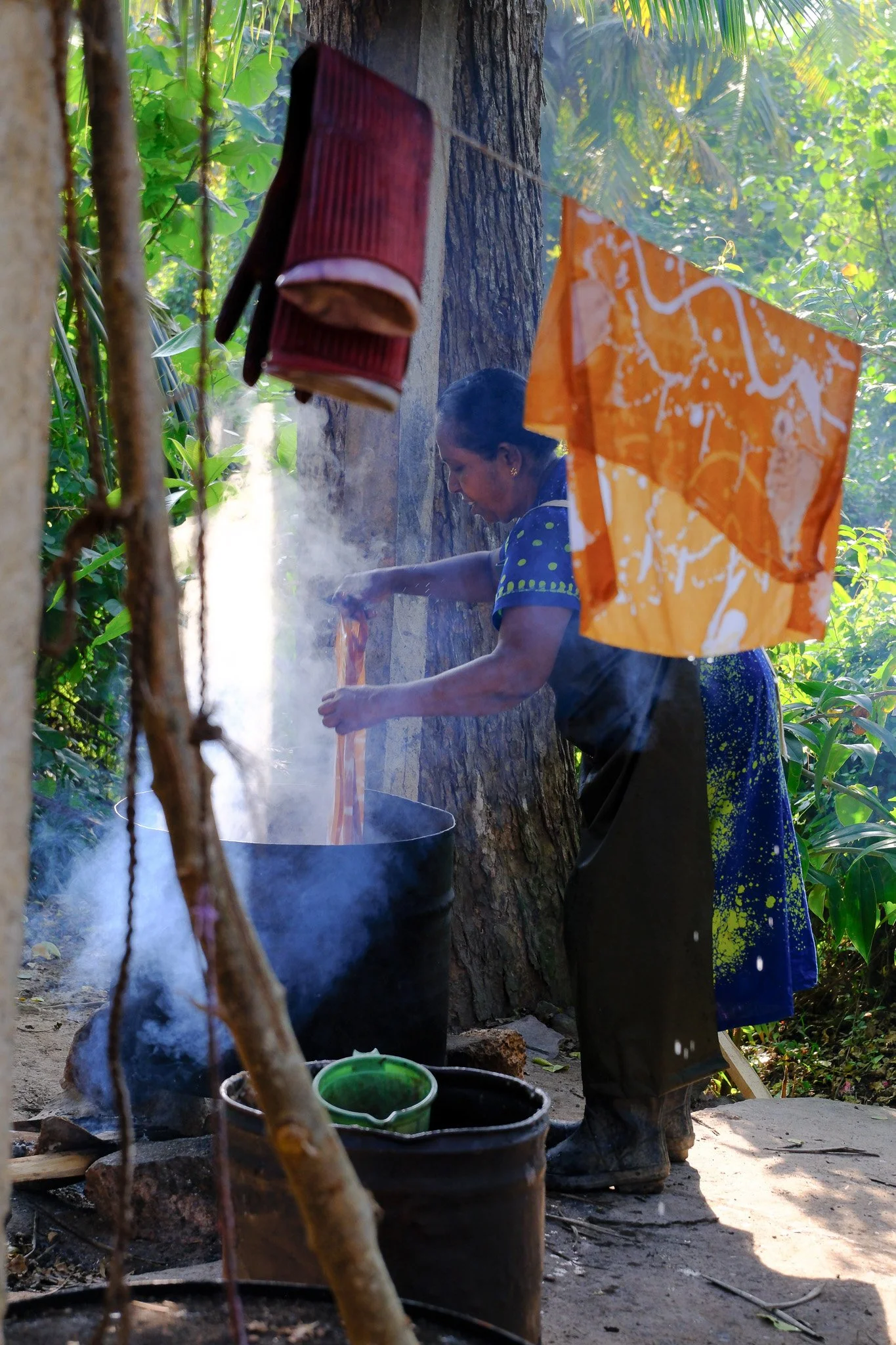 Eine Frau kocht draußen in der Natur, umgeben von grünen Pflanzen, bei Sonnenlicht.