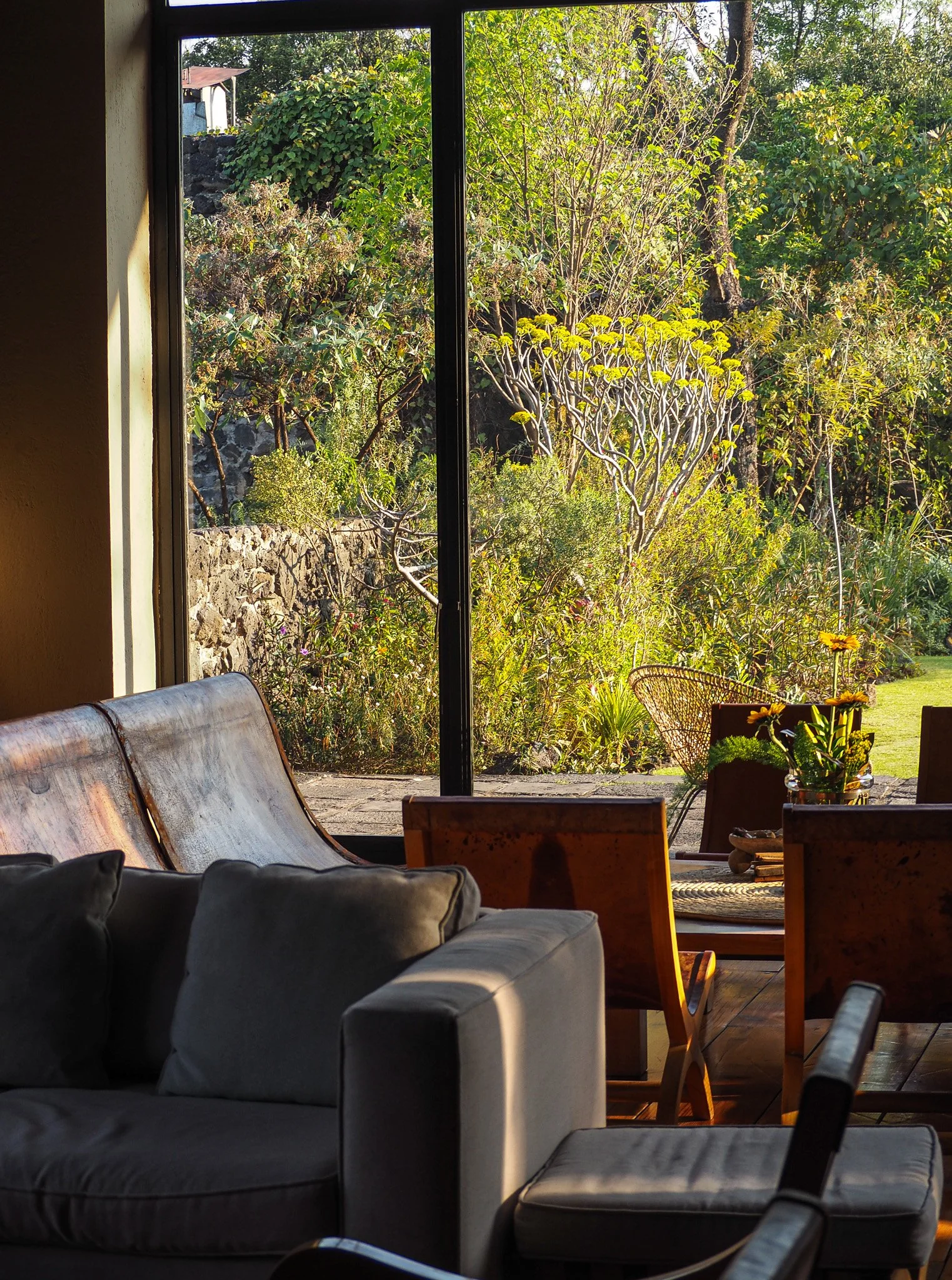 Innenraum mit einem grauen Sofa und einem Holztisch, blickt auf einen Garten mit Büschen, Bäumen und Sonnenlicht durch ein großes Fenster.