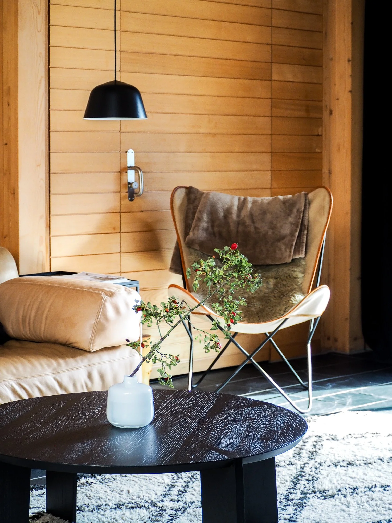 Wohnzimmer mit Holzwand, schwarzer Deckenlampe, beige Ledercouch, dekorativem Ast in weißer Vase auf Schwarzem Couchtisch, Sessel mit Mossfellen