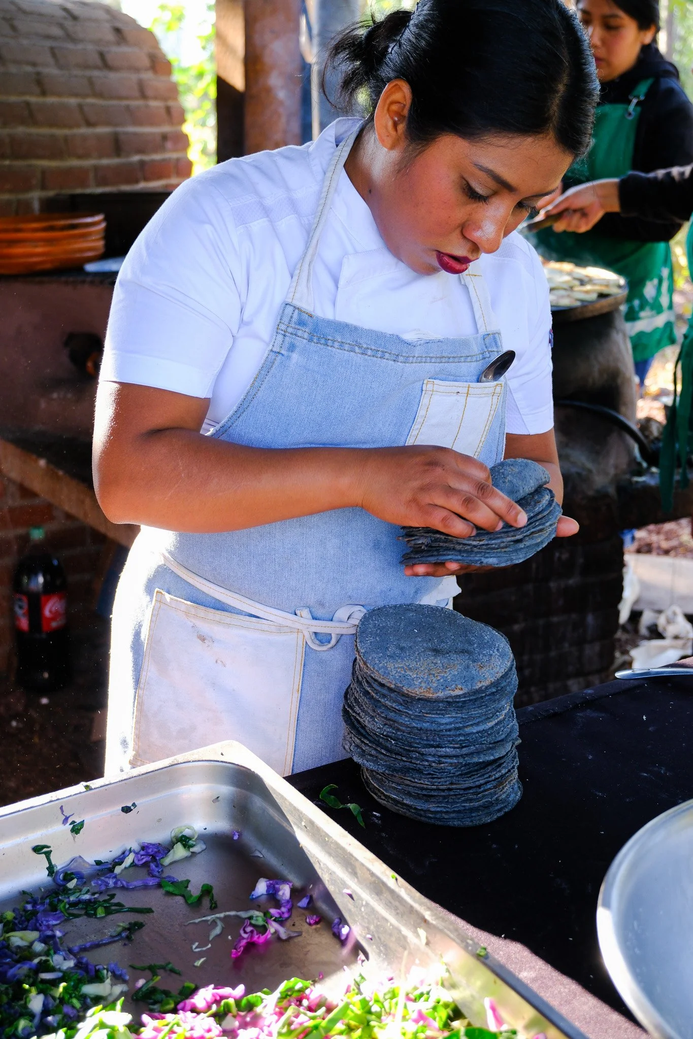 Eine Frau in Weiß mit blauer Schürze stapelt schwarze Mais-Tortillas in einem offenen Küchenbereich im Freien, im Hintergrund weitere Personen beim Kochen, mit bunten Blümchen auf einem Metallblech im Vordergrund.