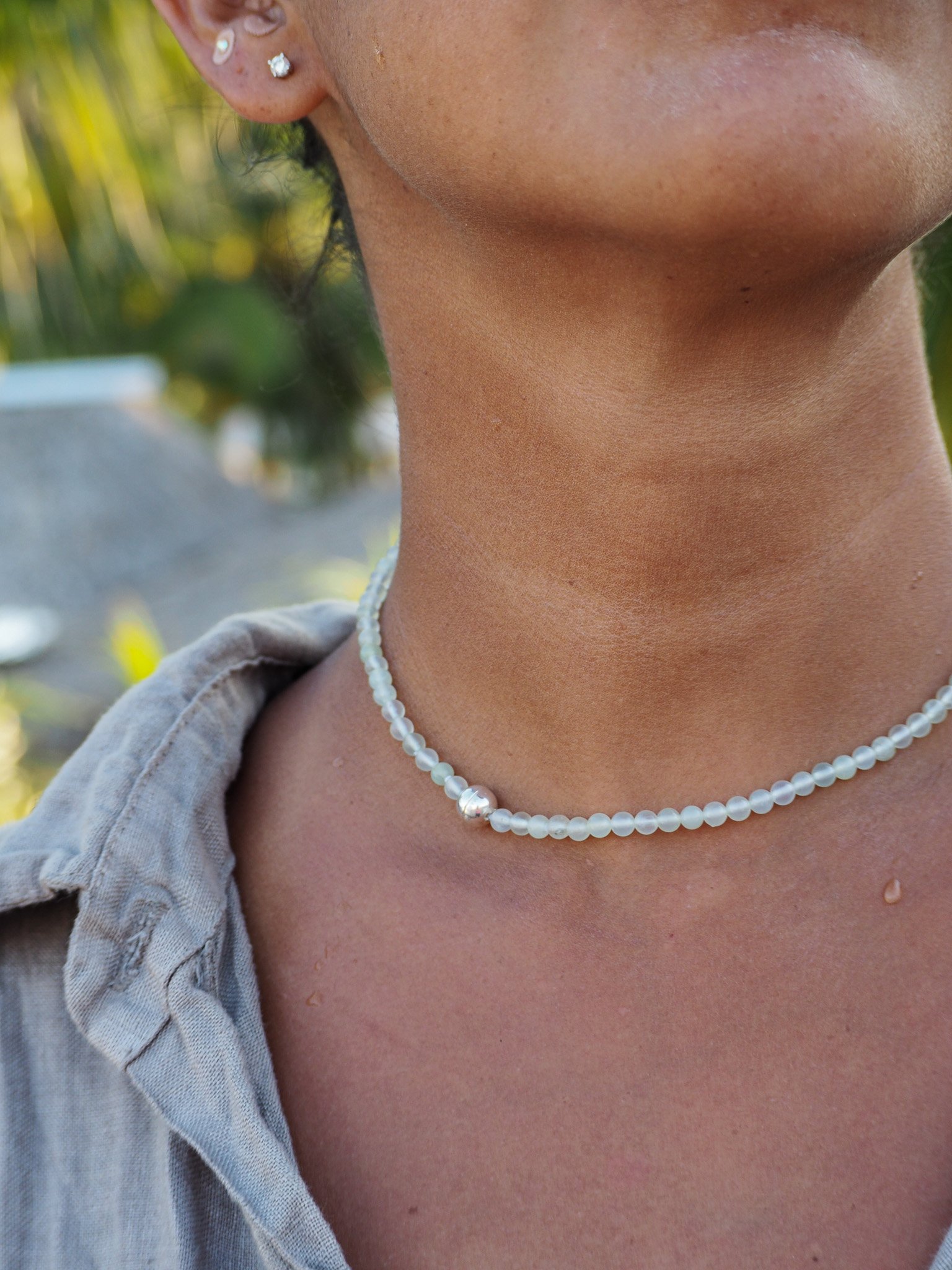 Nahaufnahme des Halses und Teils des Gesichts einer Person mit dunkler Haut, tragend eine Perlenschmuckkette und mehrere Ohrringe, vor einem natürlichen, grünen Hintergrund.