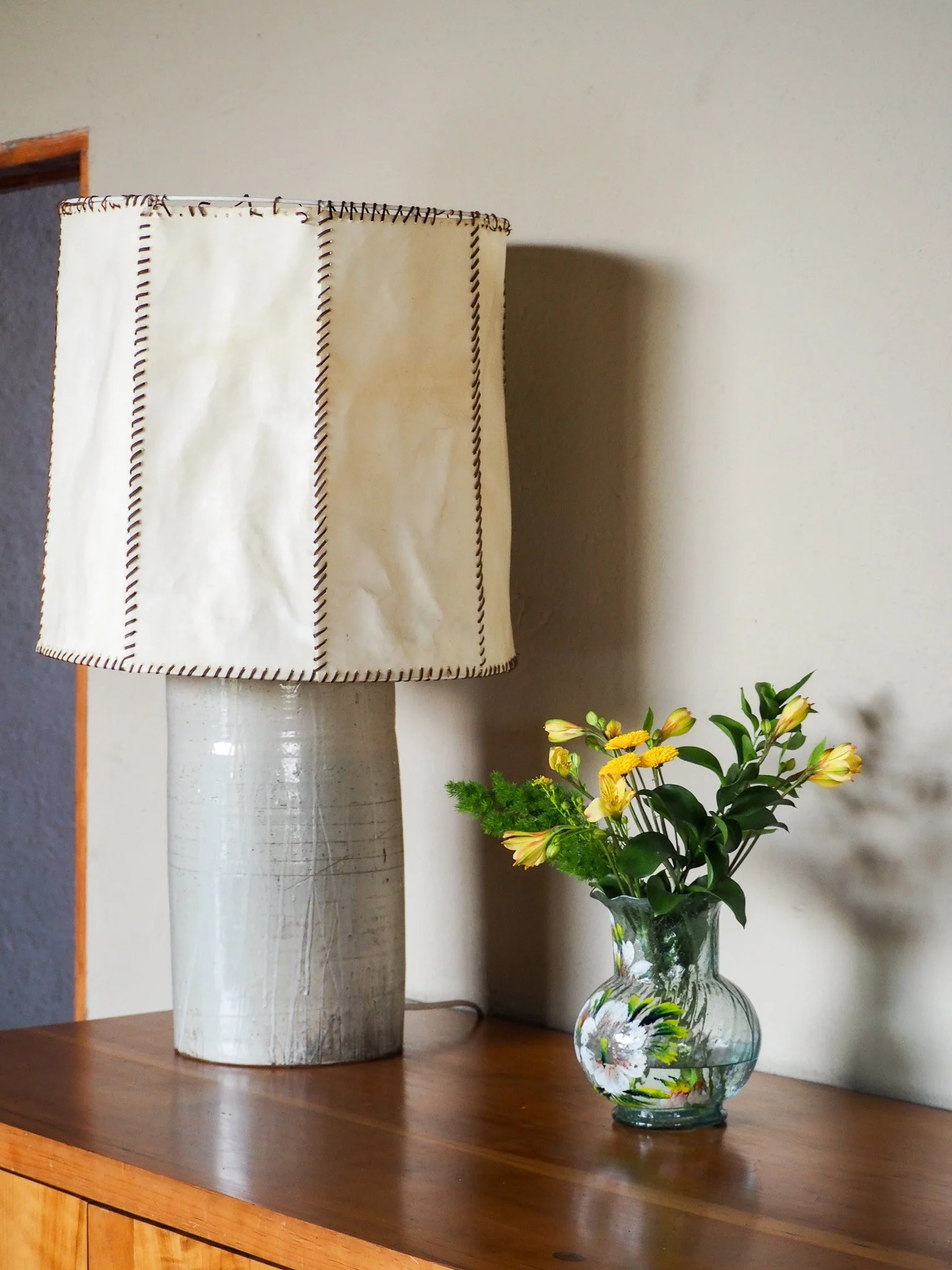 Eine Stehlampe mit einem weißen Lampenschirm neben einer Glasvase mit gelben Blumen auf einem Holztisch.