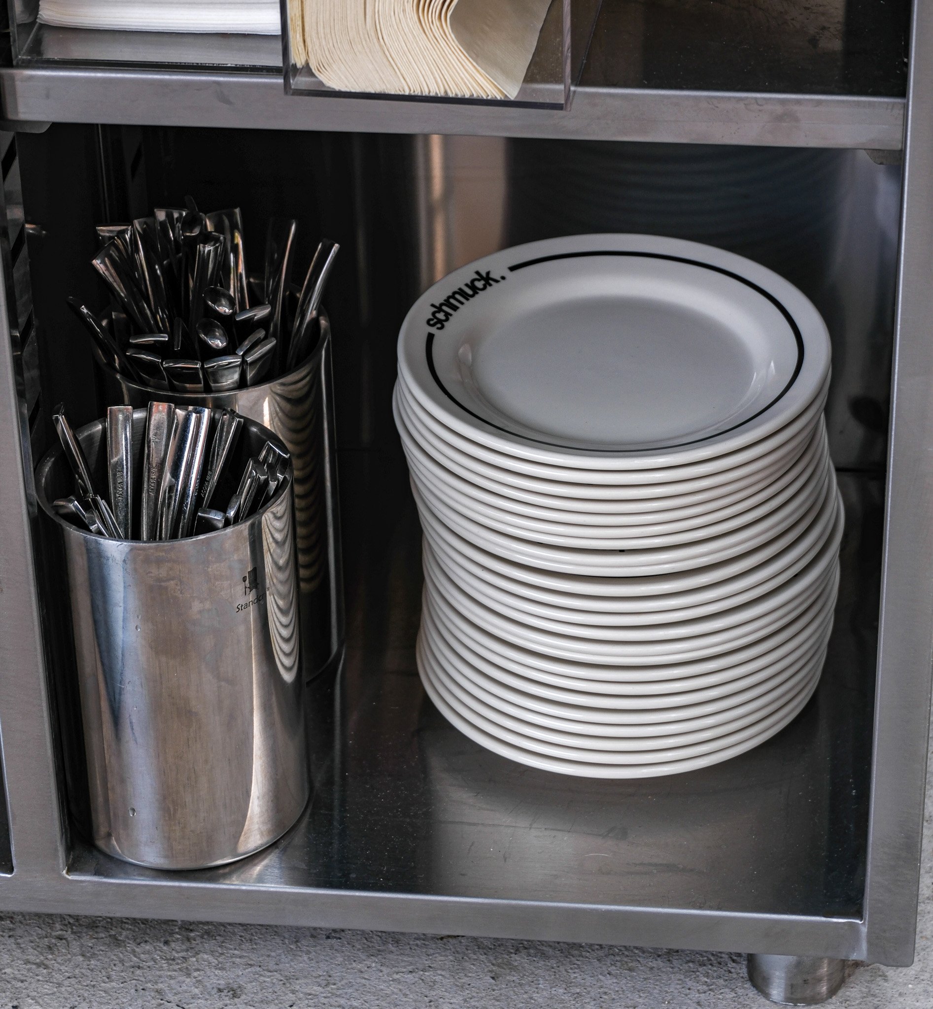 Stapel weißer Teller mit schwarzem Text und schwarzen Kreisen, Edelstahlbehälter mit Messer und Gabeln, Holzlöffel auf oberer Ablage