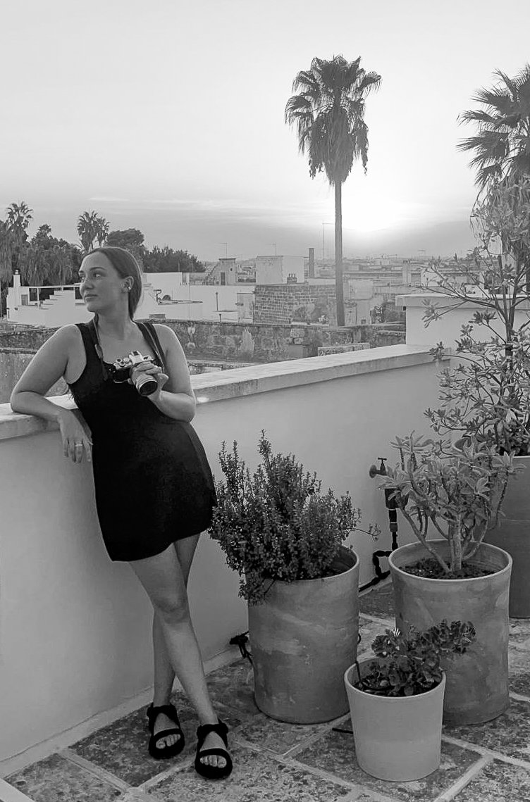 Frau mit Kamera auf einem Balkon, umgeben von Pflanzen, bei Sonnenuntergang oder Sonnenaufgang, in Schwarz-Weiß