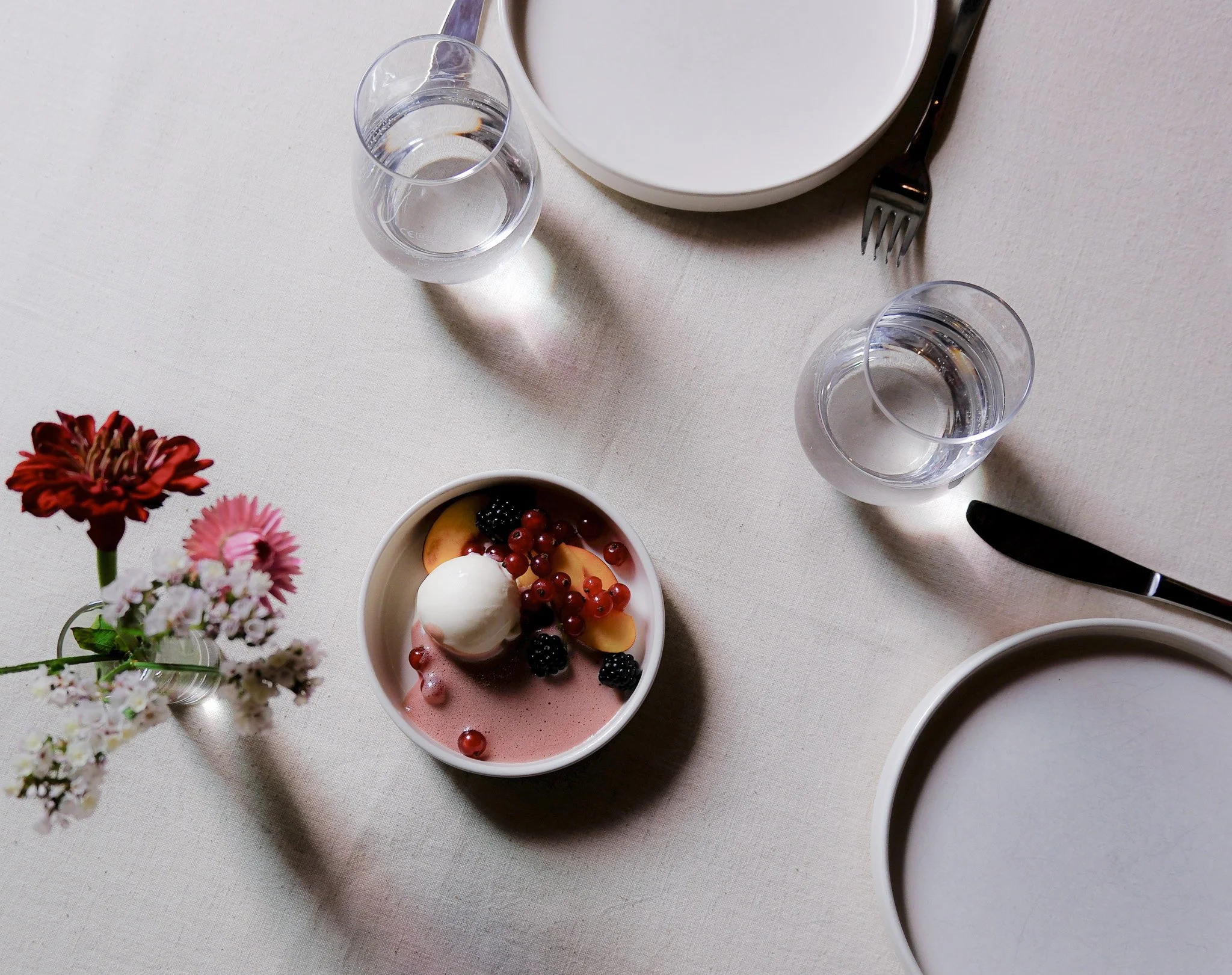 Ein gedeckter Tisch mit weißen Tellern, zwei Gläser Wasser, ein Messer, eine kleine Vase mit Blumen und eine Schale mit Dessert, das Früchte und eine Kugel Eis enthält.