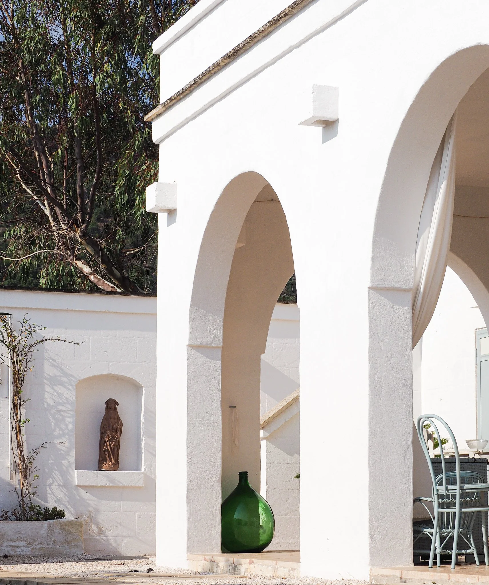 Ein weißes Gebäude mit großen offenen Bögen, einer grünen Vase und einem Sitzbereich mit Stühlen im Außenbereich.