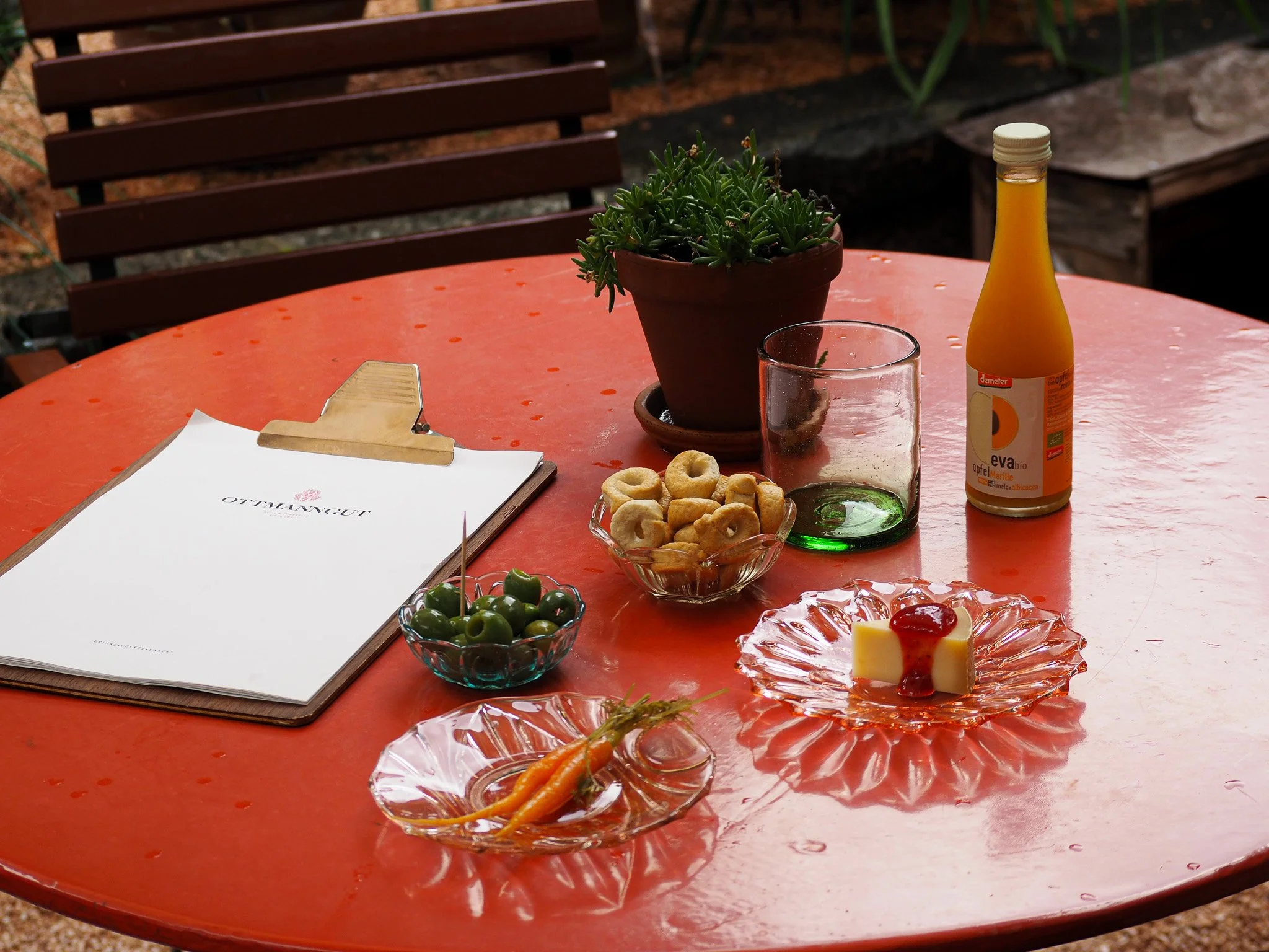 Ein runder, roter Tisch im Freien mit einer Pflanze in einem Tontopf, einem leeren Glas, einer Flasche Apfelsaft, einer Schale mit grünen Oliven, einer Schüssel mit Zwieback, einer kleinen Glasplatte mit Gürkchen, einer Schüssel mit Karotten und eine