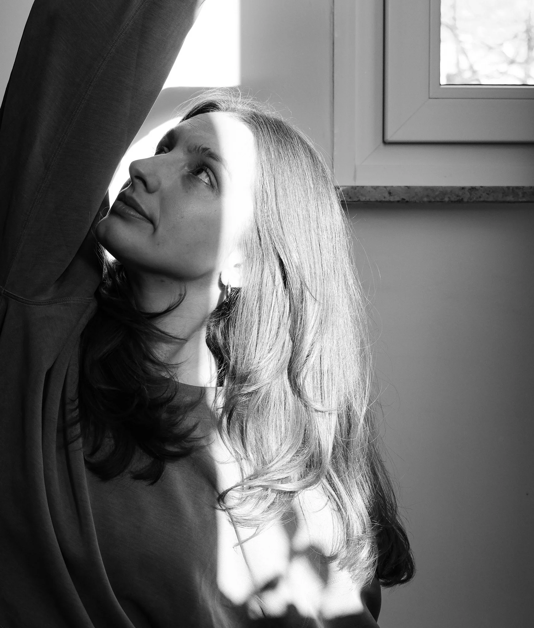 Schwarz-weiß-Foto eines jungen Mädchens mit langen, welligen Haaren, das zur Seite blickt, vor einer Fensterwand sitzt, Sonnenlicht fällt auf ihr Gesicht.