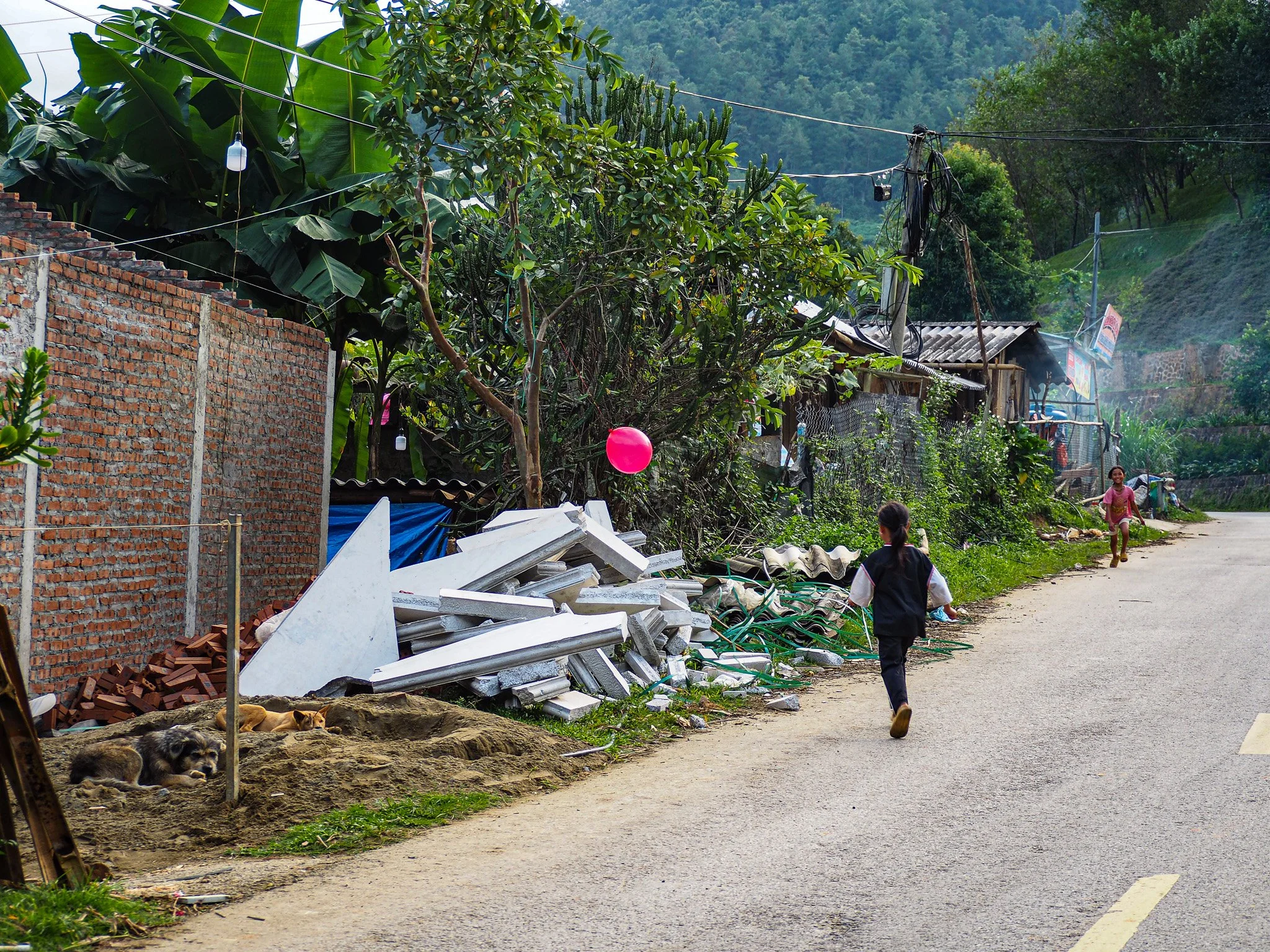 Kinder laufen auf einer ländlichen Straße, im Hintergrund Hütten, Vegetation und eine Bergkulisse, rechts liegen Baumaterialien und ein Hund ruht am Straßenrand.