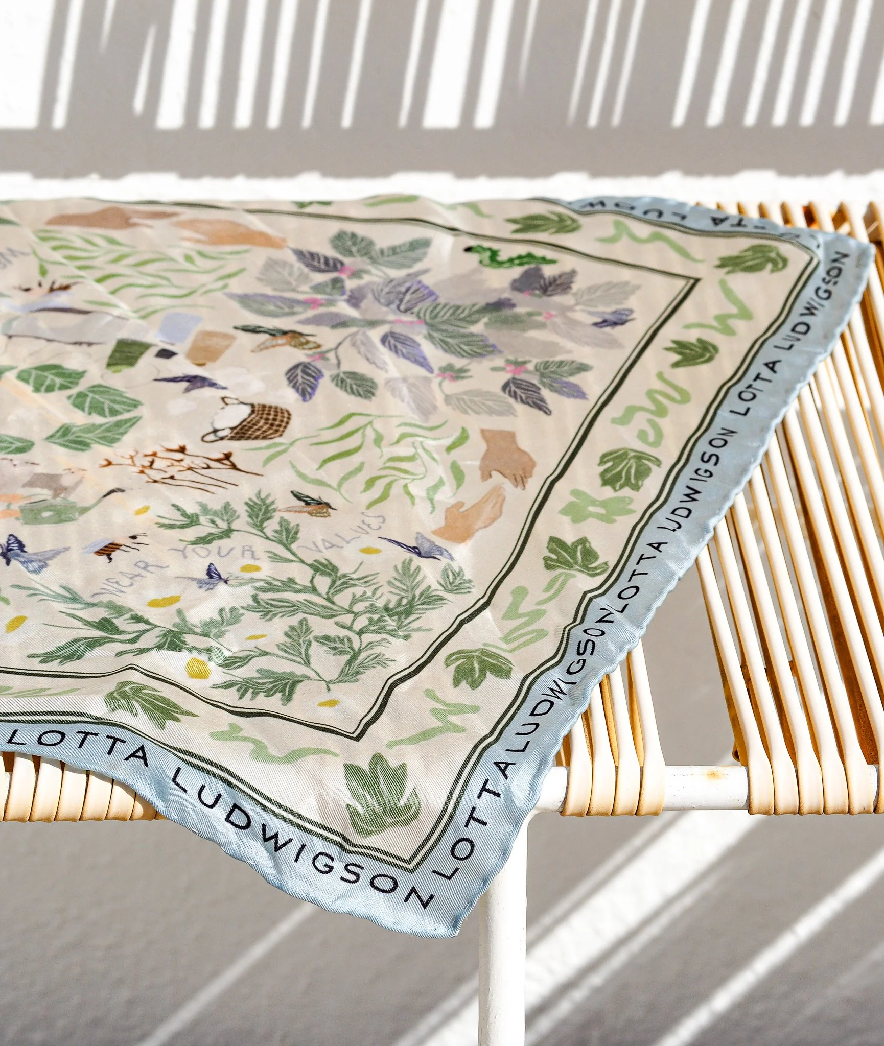 Bunte Stoffserviette mit zahlreichen botanischen Illustrationen, Blumen und Schmetterlingen, auf einem Rattantisch, mit Textrand.