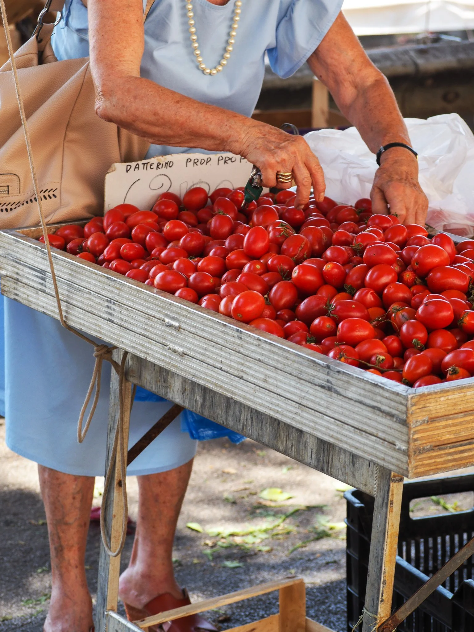 Nahaufnahme einer älteren Frau beim Tomatenverkauf auf einem Markt. Sie sortiert rote Tomaten in einem Holzstand. Sie trägt eine blaue Bluse, eine Perlenkette und mehrere Ringe.