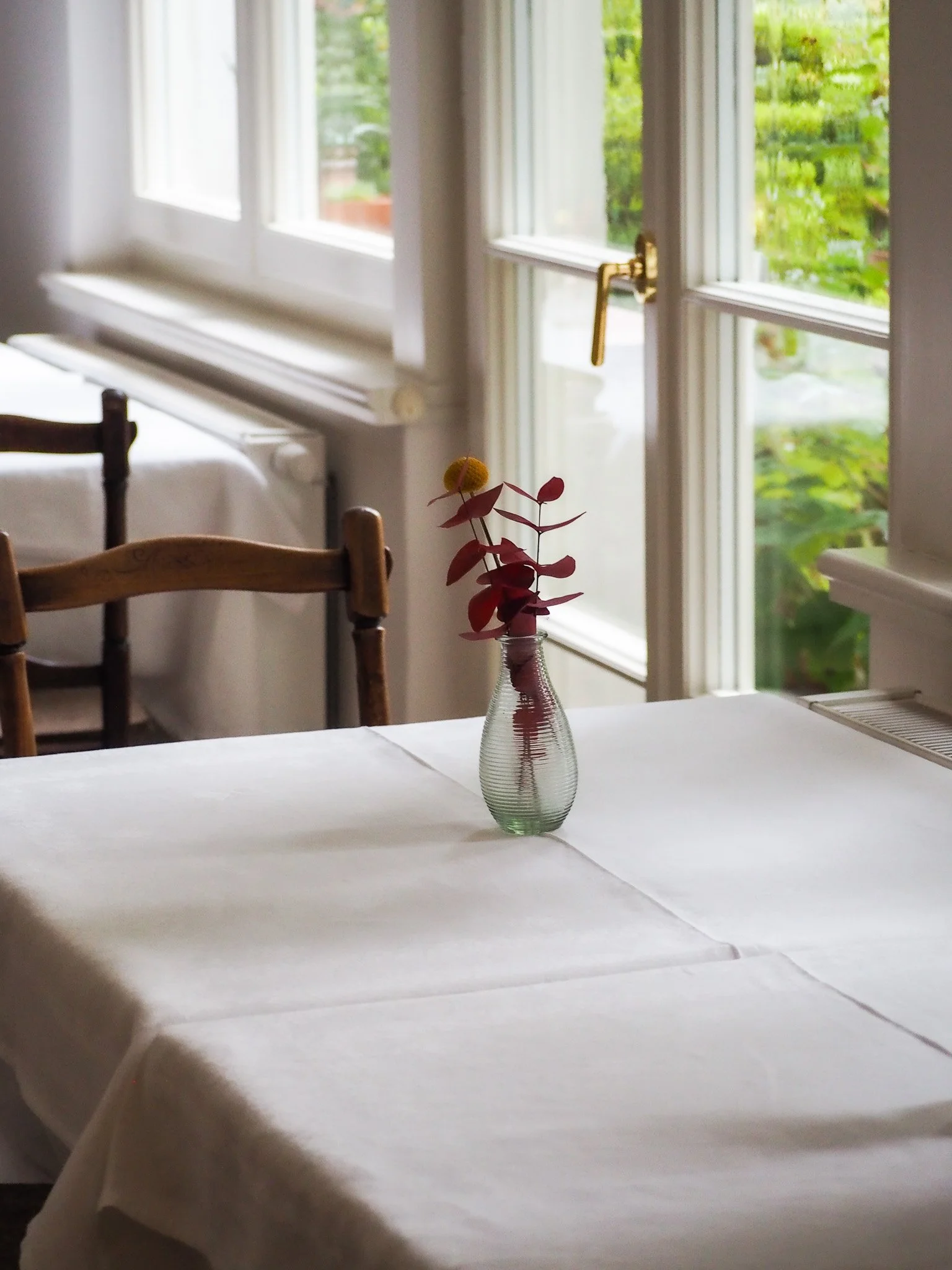 Ein weiß gedeckter Tisch mit einer kleinen Vase mit roten Blättern und einer gelben Blume, vor einem Fenster mit grüner Aussicht.