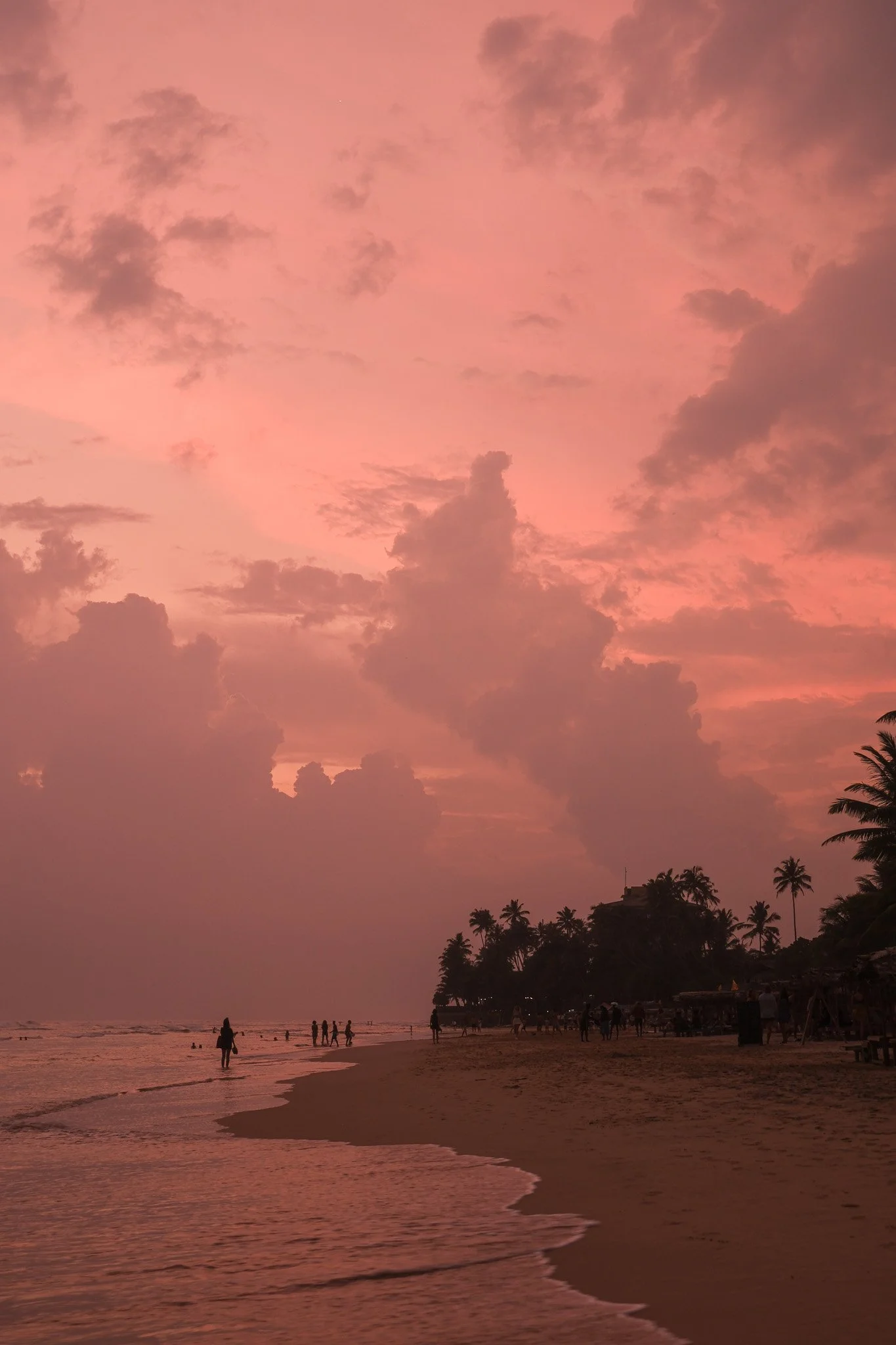 Sonnenuntergang am Strand mit pinkem Himmel und Silhouetten von Menschen und Palmen.
