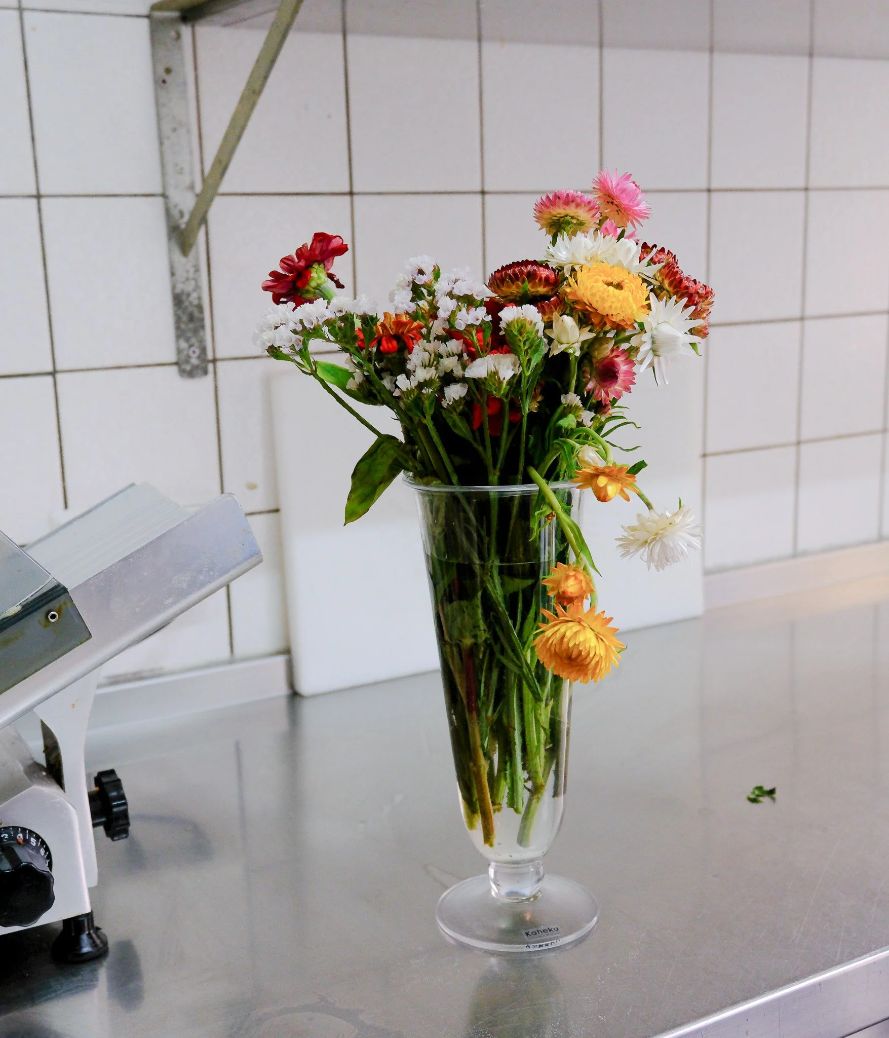 Verschiedene bunte Blumen in einer langen, klaren Vase auf einer Küchenarbeitsplatte, im Hintergrund weiße Fliesenwand.