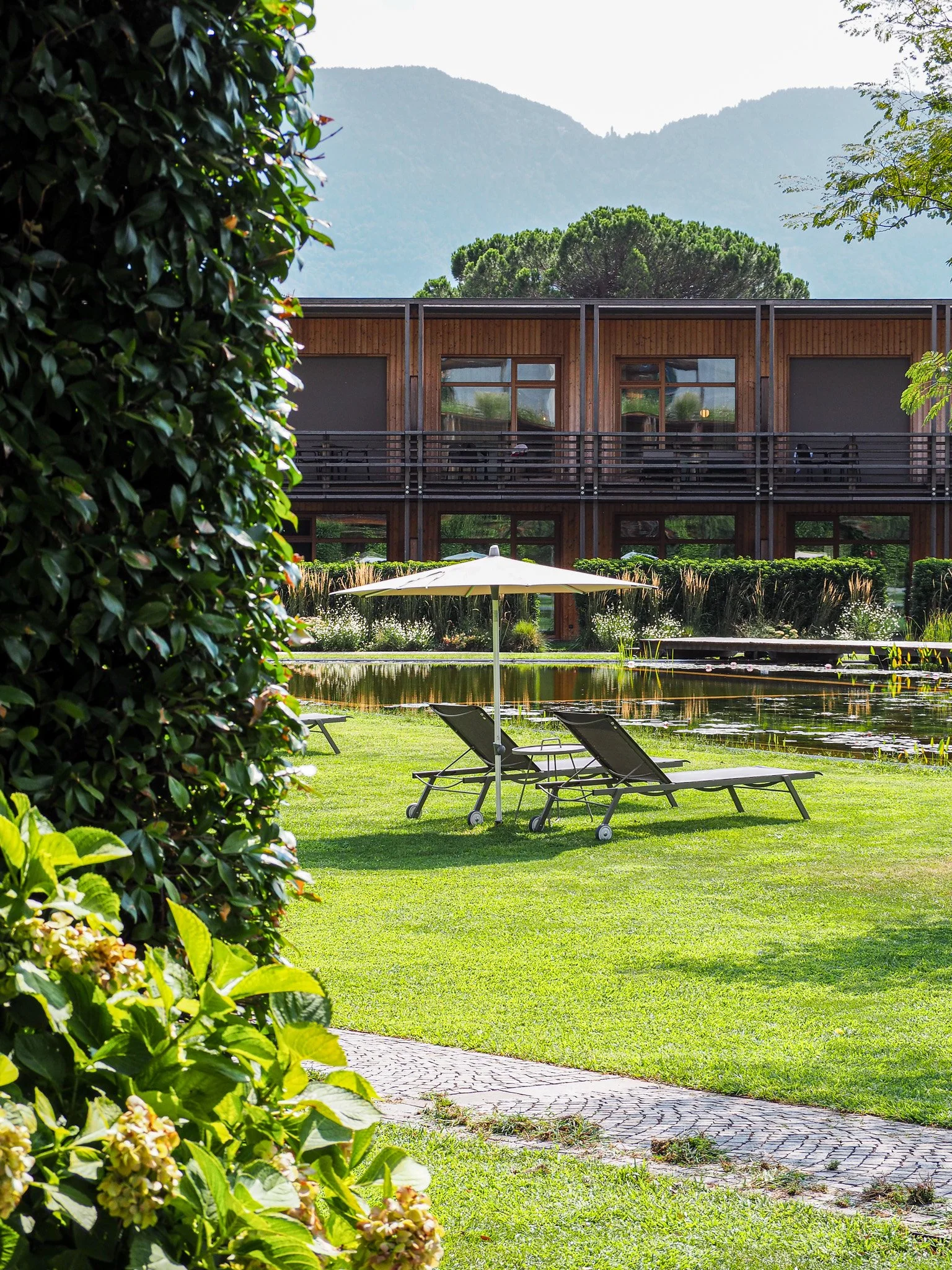 Garten mit zwei Liegestühlen und Sonnenschirm vor einem Wasserbecken, im Hintergrund ein modernes Holzgebäude, umgeben von Bäumen und Bergen.