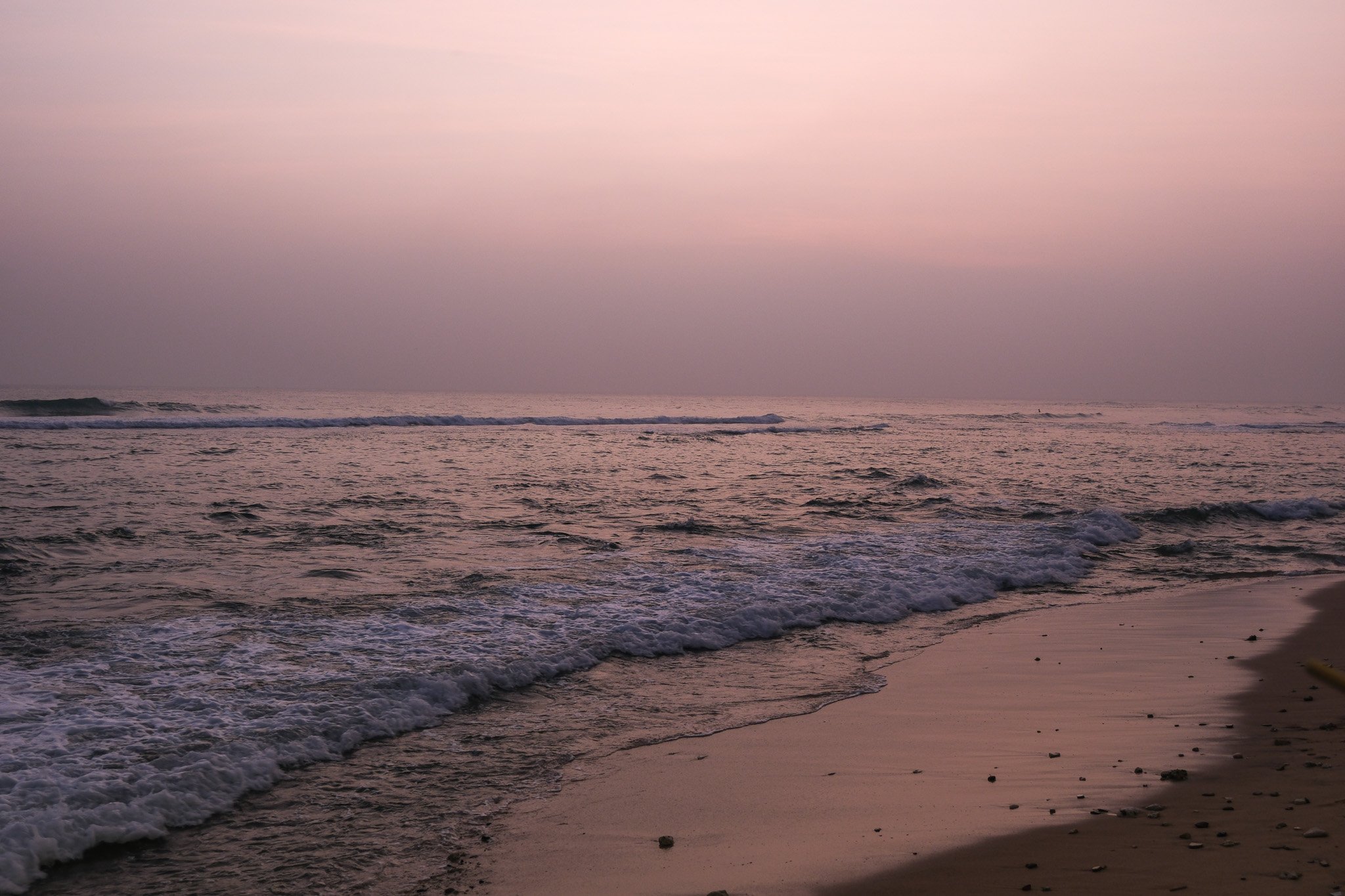 Strand bei Sonnenuntergang mit ruhigen Wellen und Sand, der mit kleinen Steinen bestreut ist.