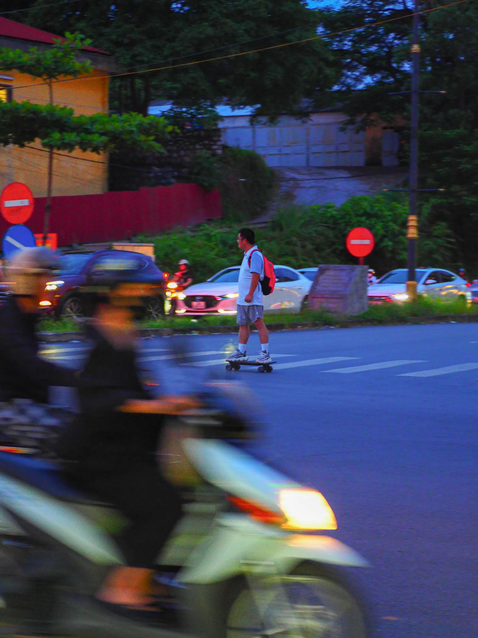 Ein Junge fährt mit einem Skateboard über die Straße, während ein Motorradfahrer vorbeifährt. Es ist Abend und im Hintergrund sind geparkte Autos und ein Zaun sichtbar.