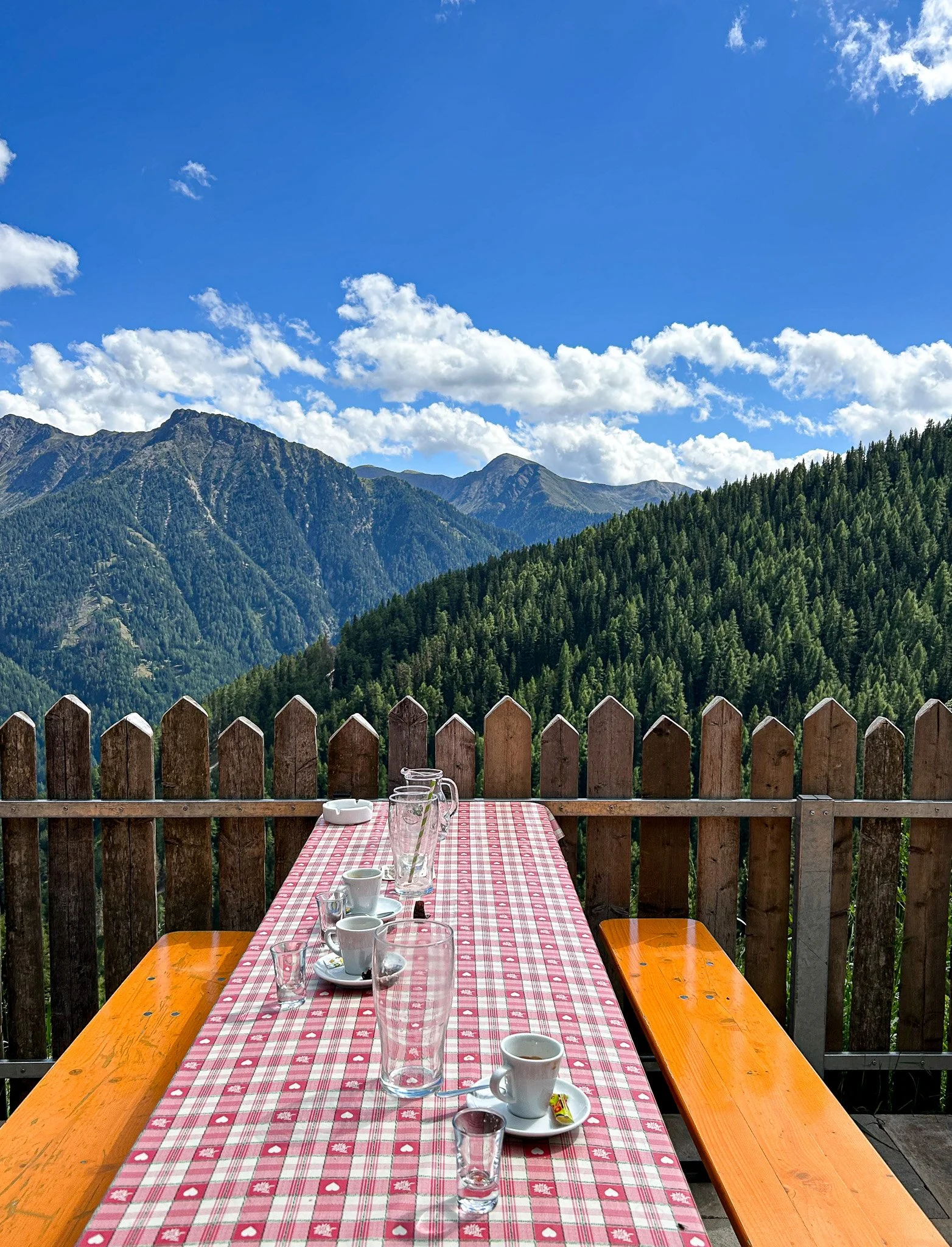 Ein Tische mit rotem Karomuster und leeren Gläsern sowie Kaffeetassen auf einer Terrasse, mit einer Holzzäunung und einer Bergkulisse im Hintergrund unter einem blauen Himmel mit weißen Wolken.
