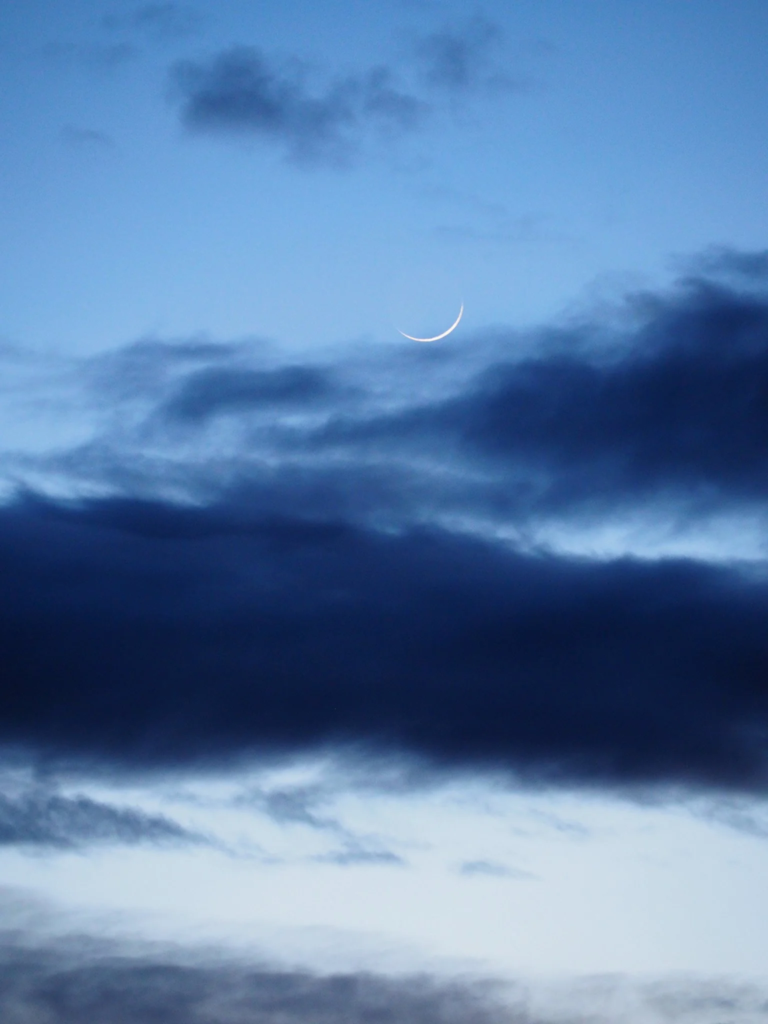 Der Himmel mit dunklen Wolken und einer schmalen Mondsichel in der Dämmerung.