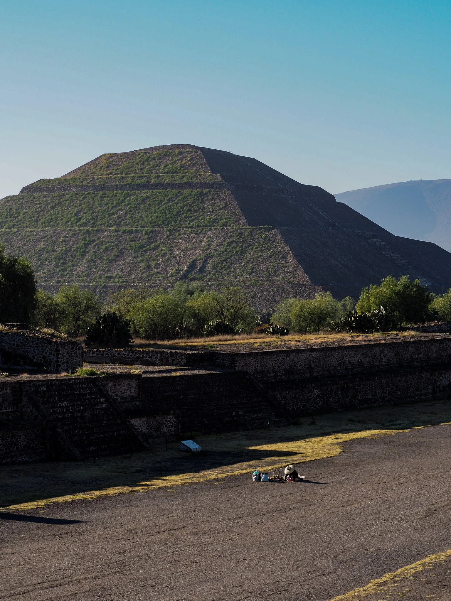Ein großer, schichtartiger Pyramiden-Tempel in der antiken Stadt Teotihuacán, umgeben von grünen Büschen und Bäumen, mit einer Person, die sich im Vordergrund auf dem Boden ausruht.