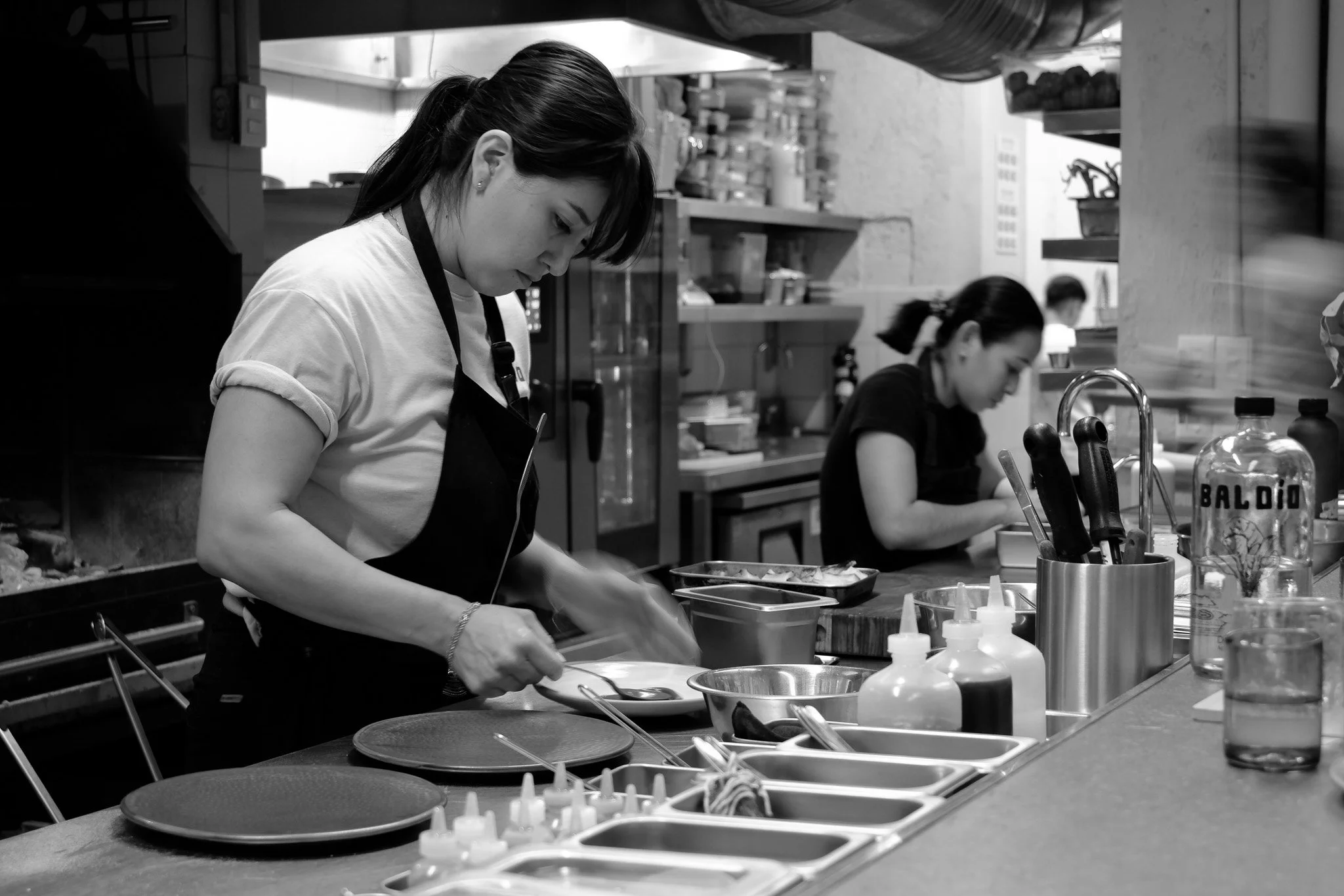 Zwei Frauen in einer Restaurantküche, eine beim Vorbereiten von Speisen, die andere im Hintergrund bei der Arbeit.