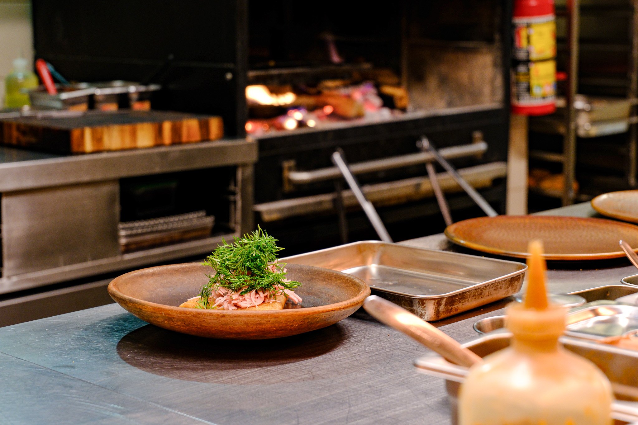 Ein unscharfes Bild einer Küche mit einem Grill im Hintergrund. Auf einem Metalltisch liegt ein Teller mit einem Gericht, garniert mit frischen Kräutern.