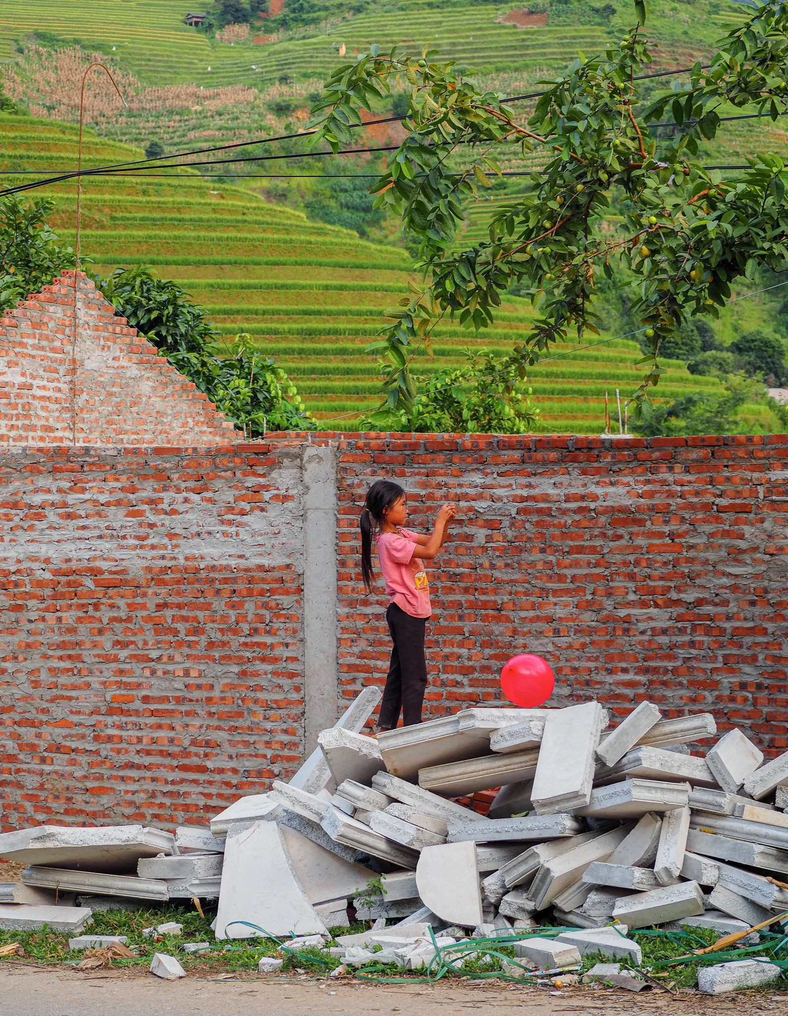 Ein Mädchen steht auf einem Stapel aus Betonteilen hinter einer Ziegelmauer und spielt mit einem roten Ball, im Hintergrund grüne Terrassenfelder und Bäume.