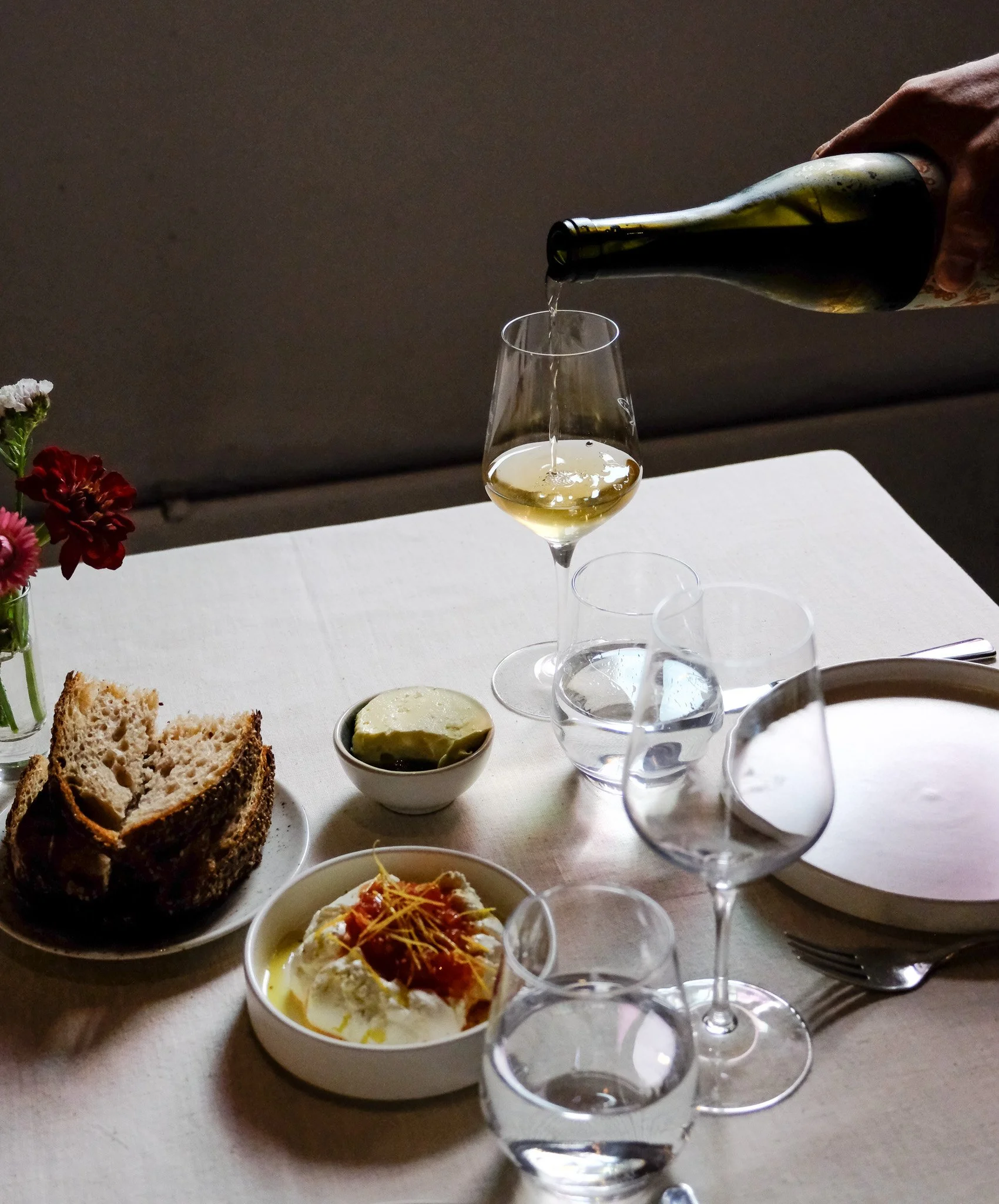 Ein gedeckter Tisch mit Wein, Wasser, Brot, Butter und einem Teller mit Käse oder Aufschnitt, zusätzlich mit einem kleinen Blumenstrauß.