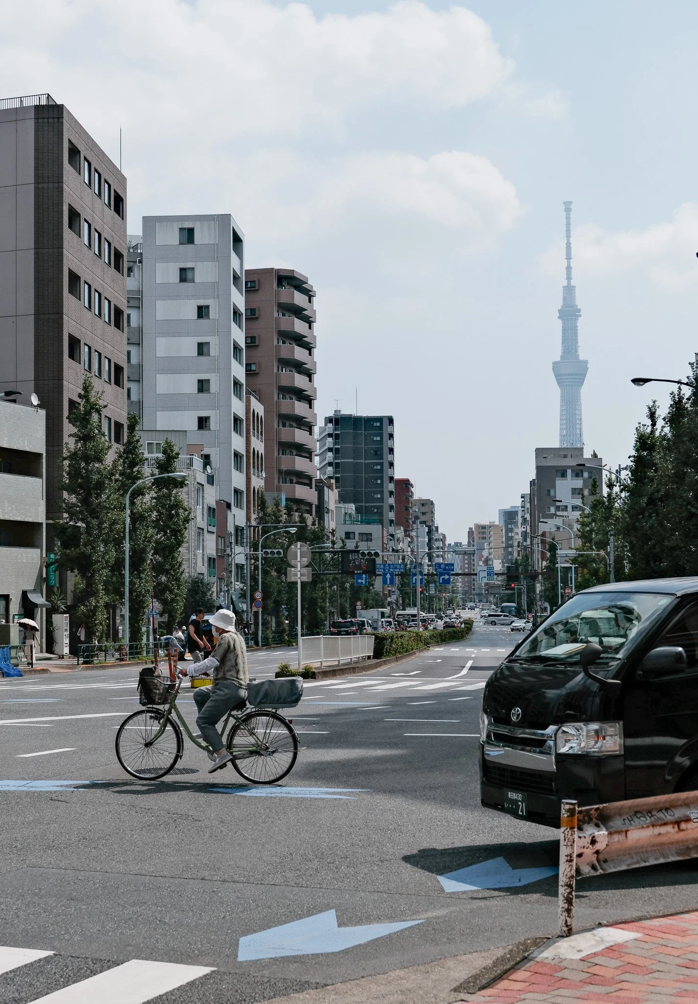 Stadtansicht mit Hochhäusern, Fahrradfahrer und Straßenverkehr, Tokyo Skytree im Hintergrund, blauer Himmel mit Wolken.