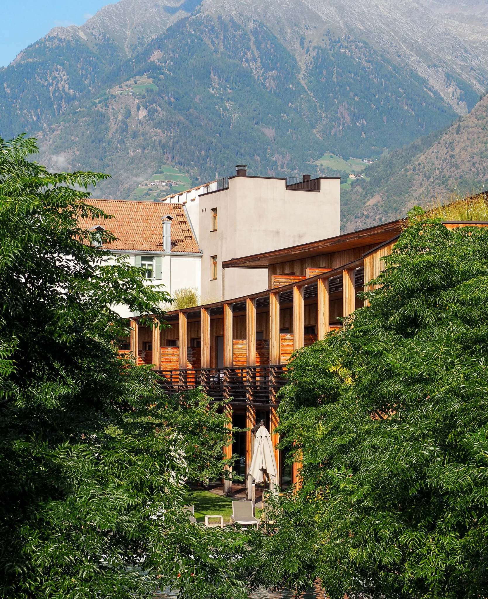 Ein modernes Gebäude mit Holzfassade und Balkon, umgeben von grünen Bäumen und Bergen im Hintergrund, bei Sonnenlicht.