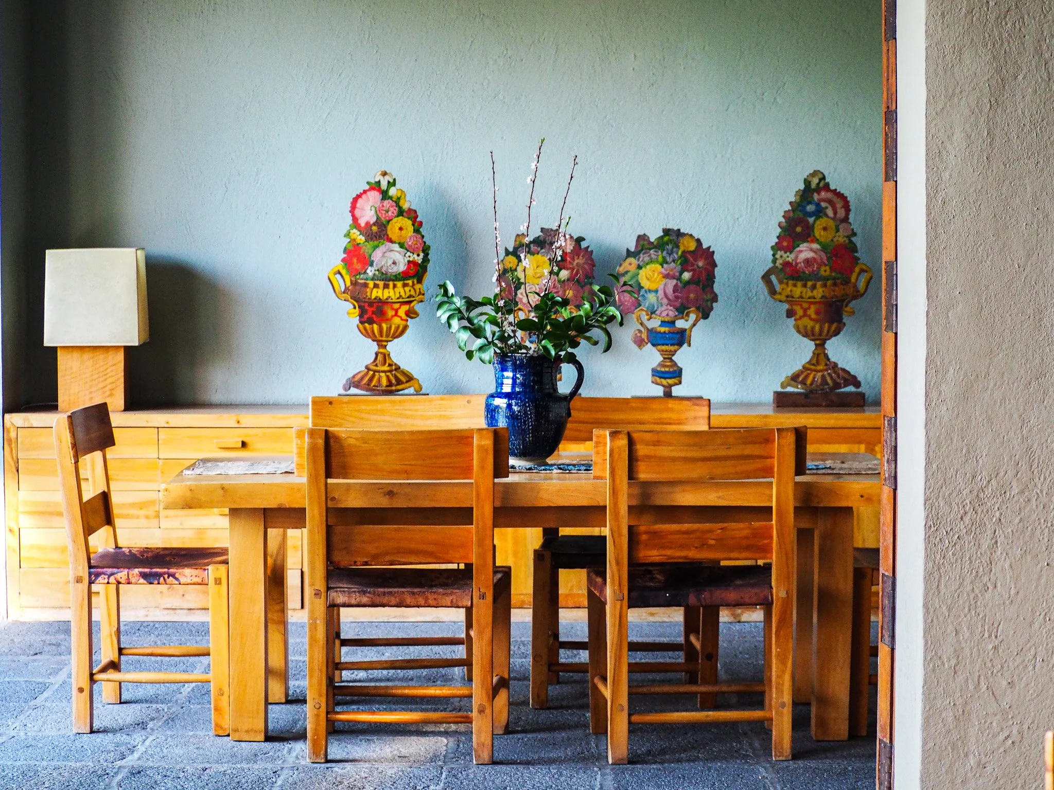 Ein Holztisch mit sechs Holzstühlen, eine dunkelblaue Vase mit Blumen in der Mitte, im Hintergrund eine verglaste Wand mit drei bunten Blumenvasen
