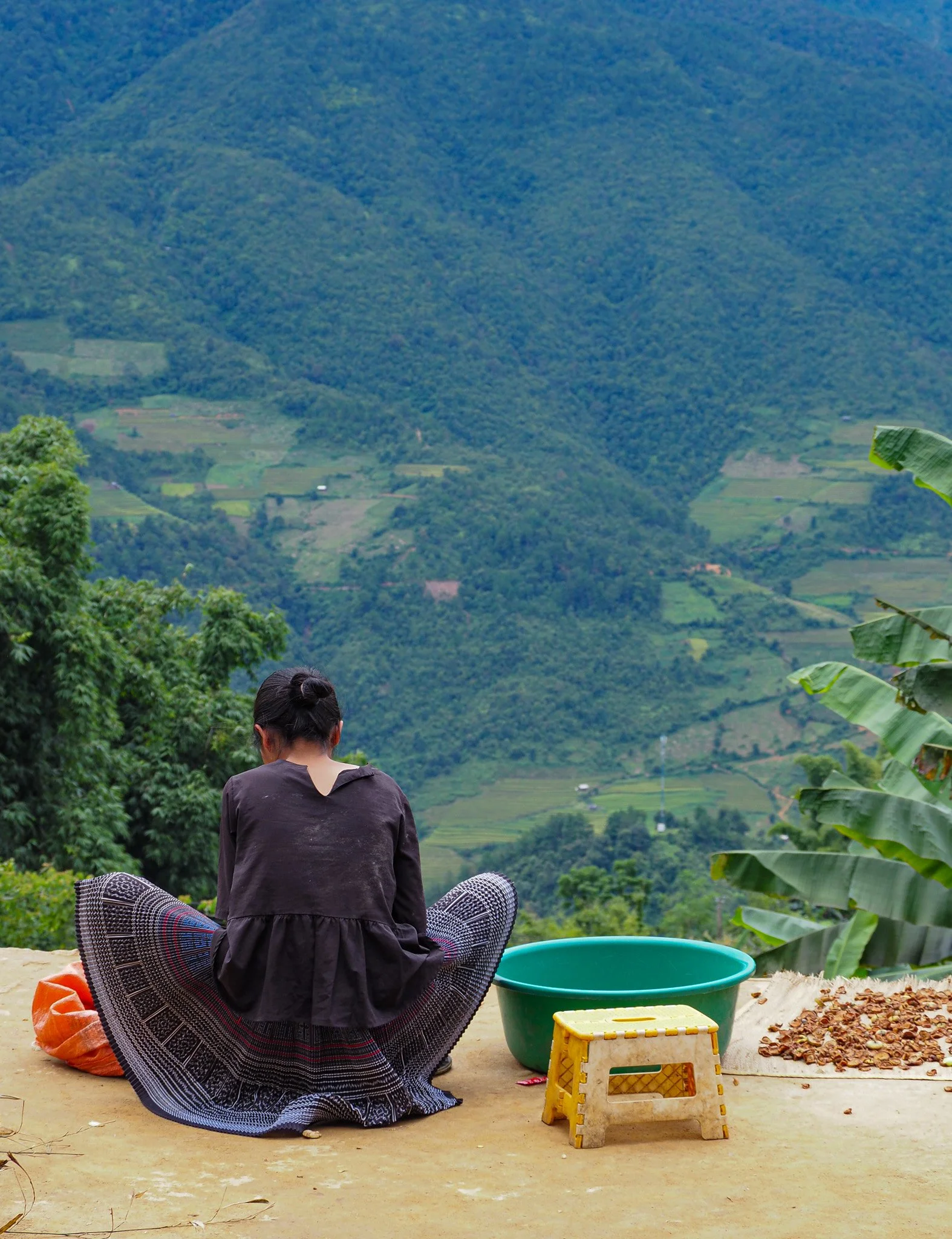 Ein Mädchen sitzt auf dem Boden mit Blick auf eine grüne Berglandschaft, umgeben von Bananenpflanzen, mit einem grünen Behälter, einer gelben Hocke und einer kleinen Menge Nussschale neben ihm.
