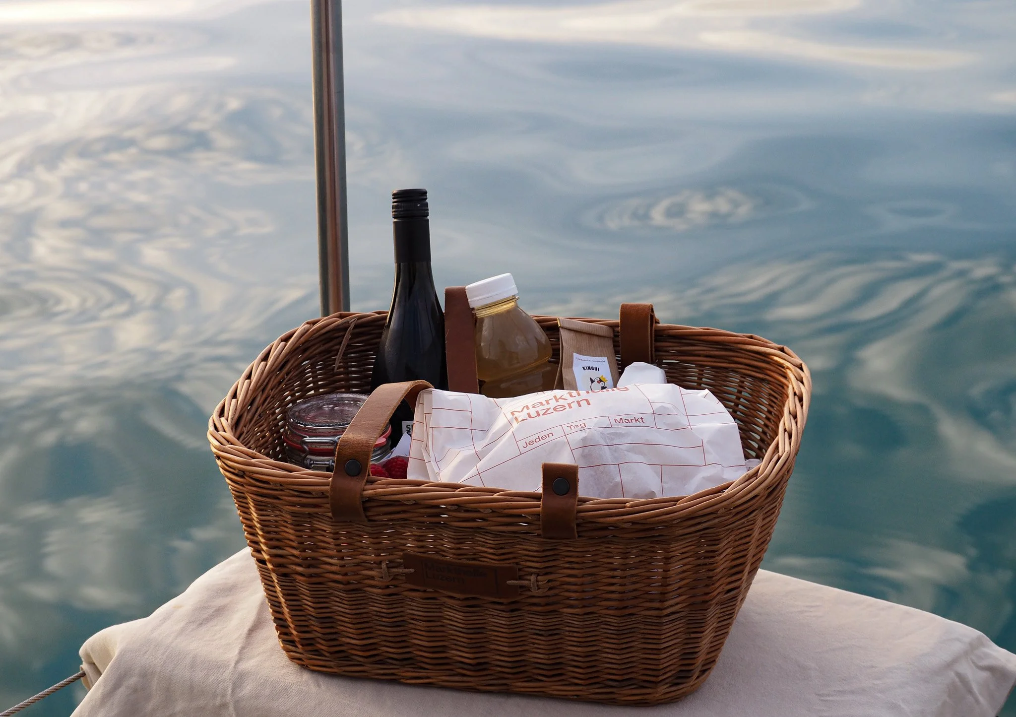 Ein Picknickkorb mit Weinflasche, Marmelade, Tüten und weiteren Lebensmitteln auf einer weißen Decke vor dem Wasser bei Sonnenuntergang.
