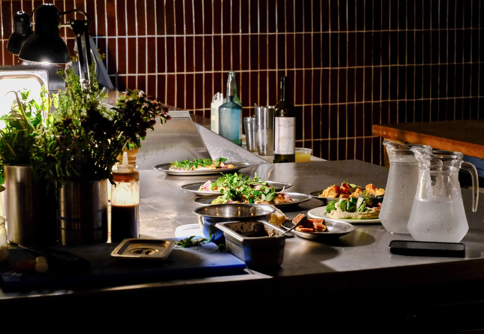 Eine mitessen bestückte Küchenarbeitsfläche mit verschiedenen Tellern, Gläsern, Wasserkrügen, Weinflasche, Kräutertöpfen und Küchenutensilien, beleuchtet von einer Anbaulampe.