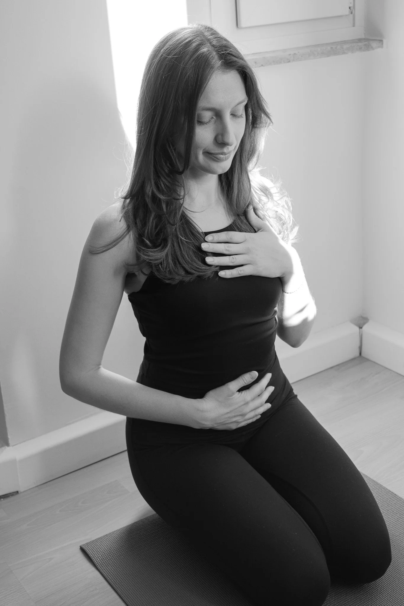 Eine Frau macht Yoga in einem Raum, kniet auf einem Yoga-Matte, mit den Händen auf Brust und Bauch, Augen geschlossen.