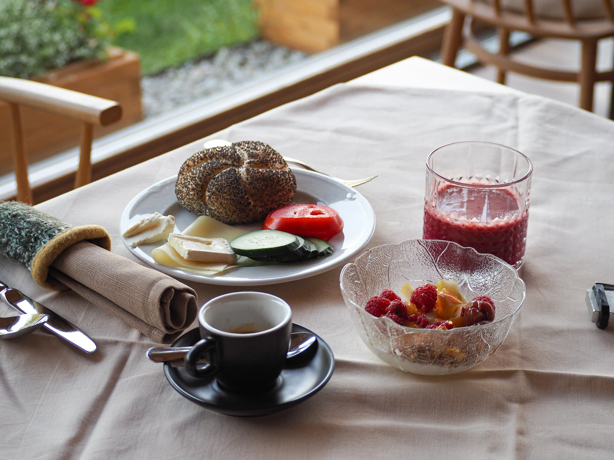 Ein Frühstückstisch mit belegten Brötchen, Kaffee, Joghurt mit Beeren, Orangensaft, Tomatenscheiben, Käse, Gurkenscheiben und ein Brotkorb, auf einem hellen Tisch in einem hell erleuchteten Raum.