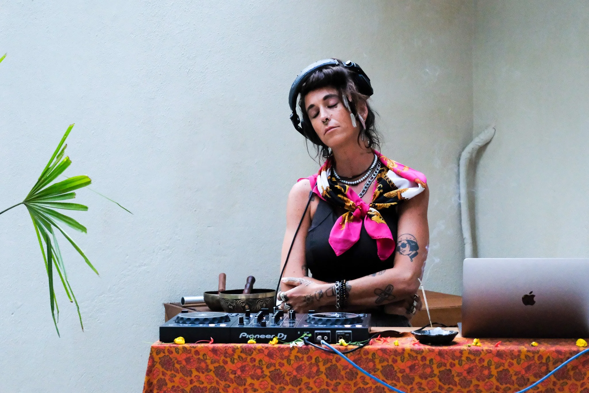 Eine weibliche DJ mit dunklen, kurzen, gewellten Haaren und mehreren Tattoos, die einen schwarzen Kopfhörer trägt, steht hinter einem DJ-Controller auf einem Tisch mit rotem, gemustertem Tuch, neben einem Laptop und einer Handglocke, in einem Raum mit schiefergrauem Wandhintergrund.