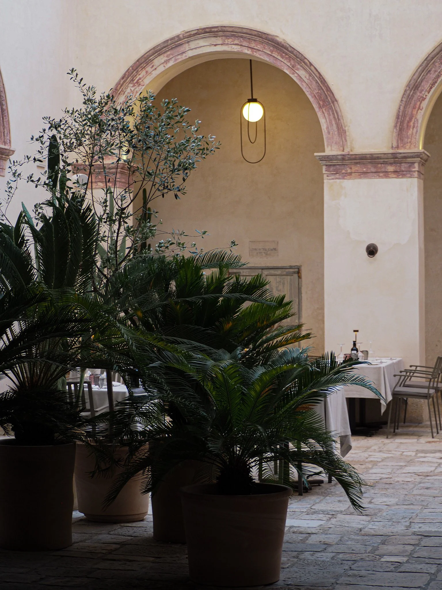 Innenraum eines alten Gebäudes mit hohen Wänden, einer im Hintergrund hängenden Lampe, mehreren großen Topfpflanzen und einem gedeckten Tisch mit Stühlen im rechten Hintergrund.