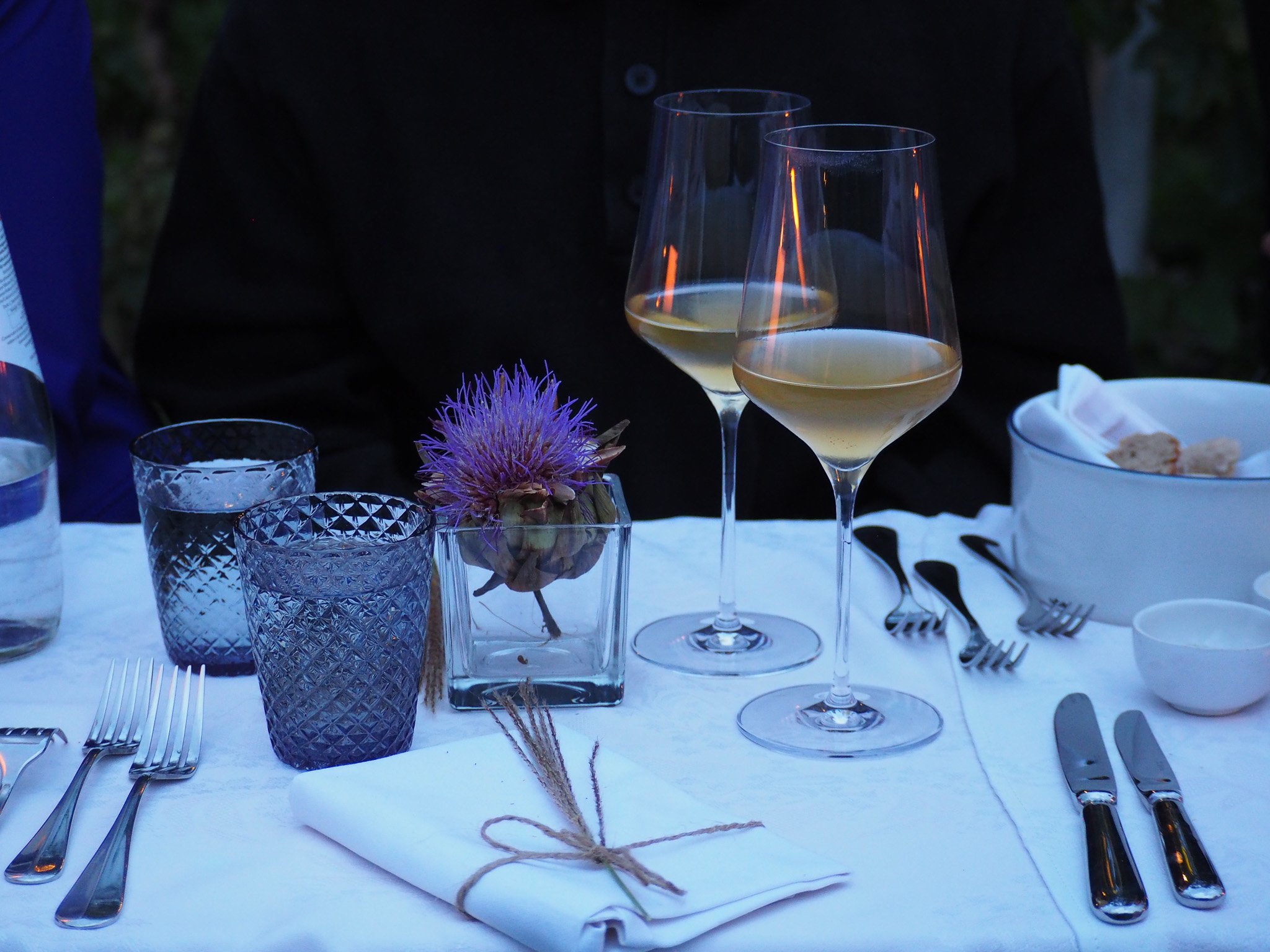 Ein gedeckter Tisch mit Weingläsern, Wasserkrügen, einer Blume in einer Vase und Geschirr, vermutlich bei einem Abendessen.