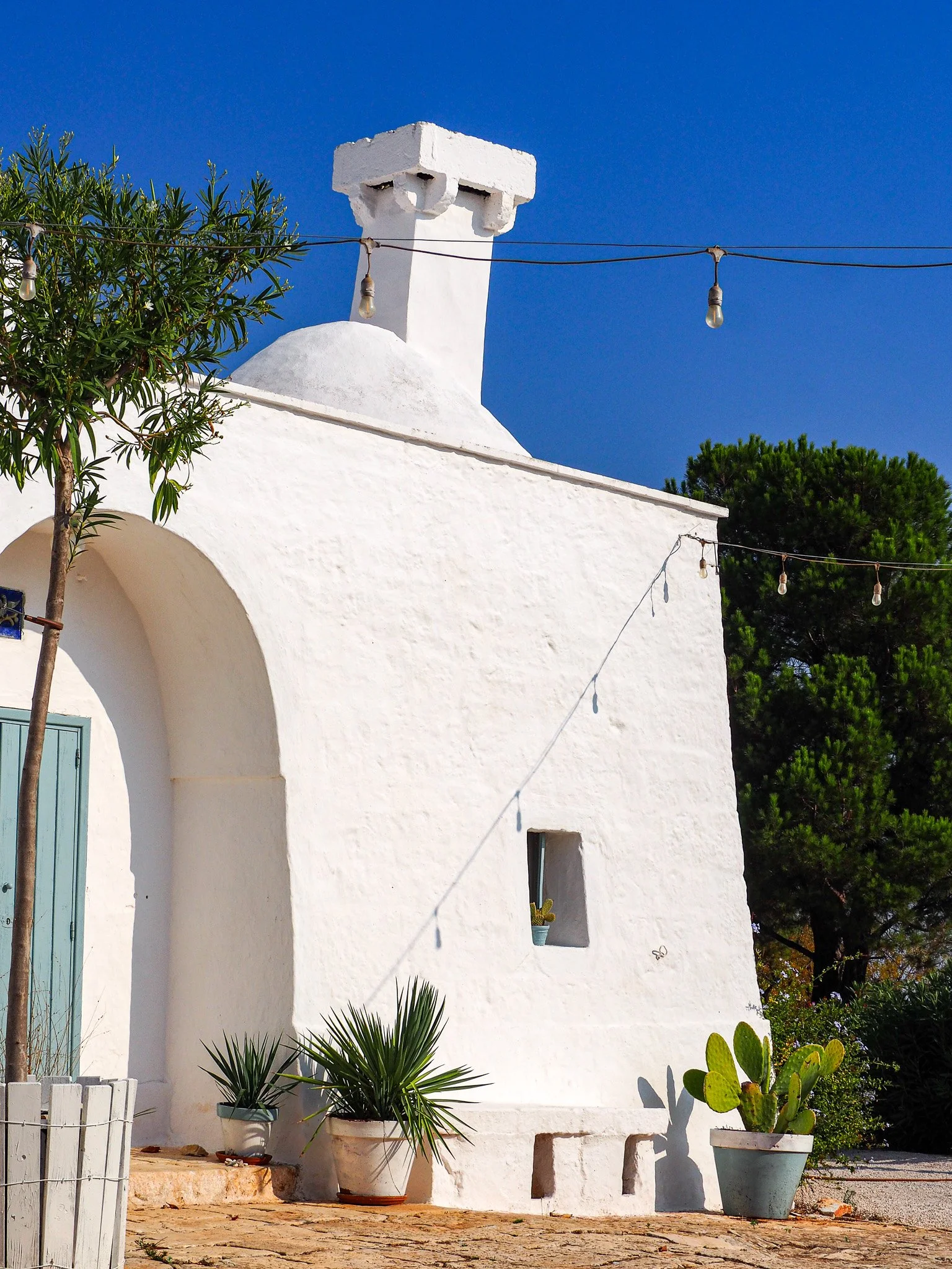 Weiße mediterrane Hausfassade mit grünen Pflanzen und Lampen gegen blauen Himmel.