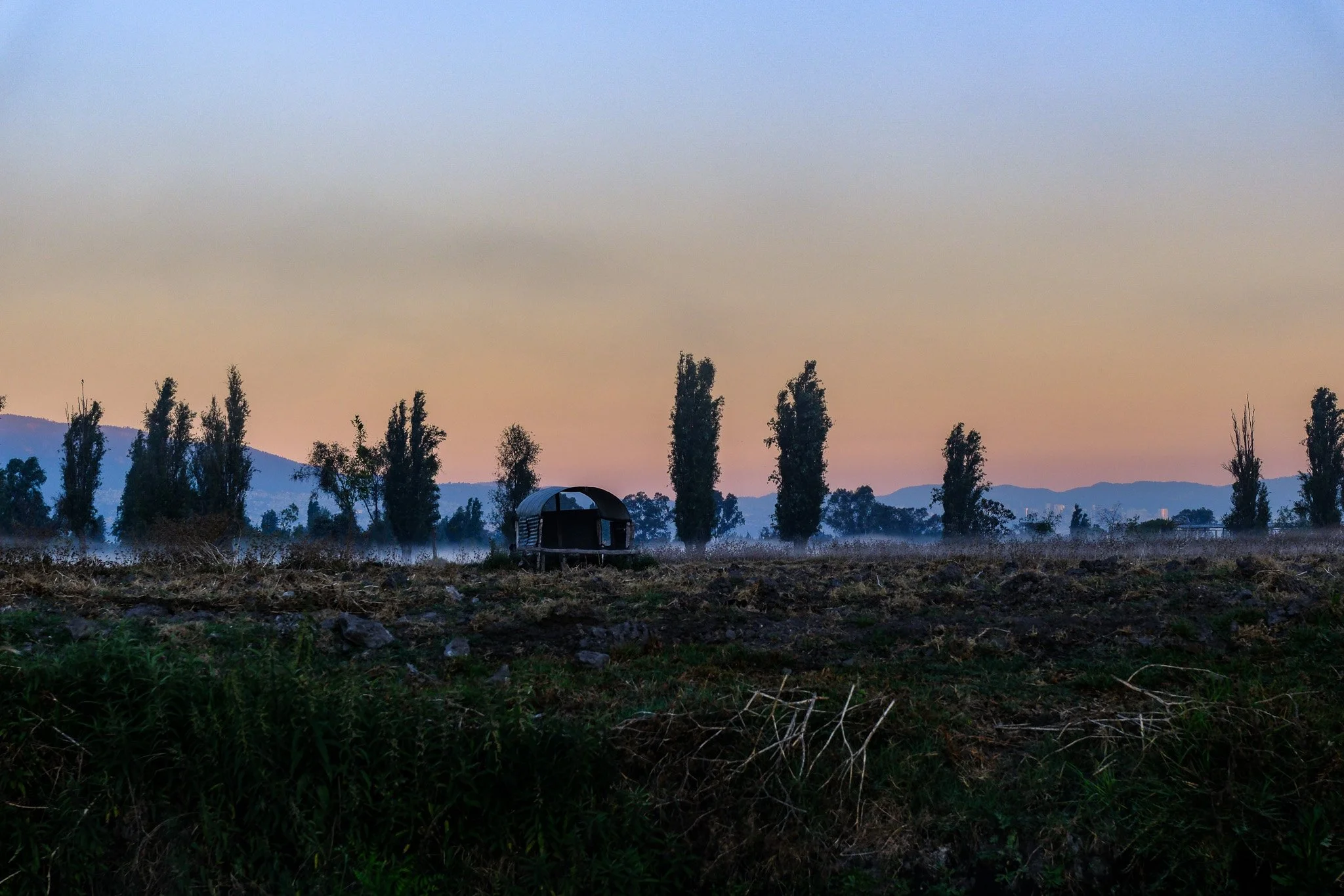 Ein kleines Haus oder Hütte in einer ländlichen Gegend bei Sonnenuntergang, umgeben von Bäumen und teilweise bewölktem Himmel.