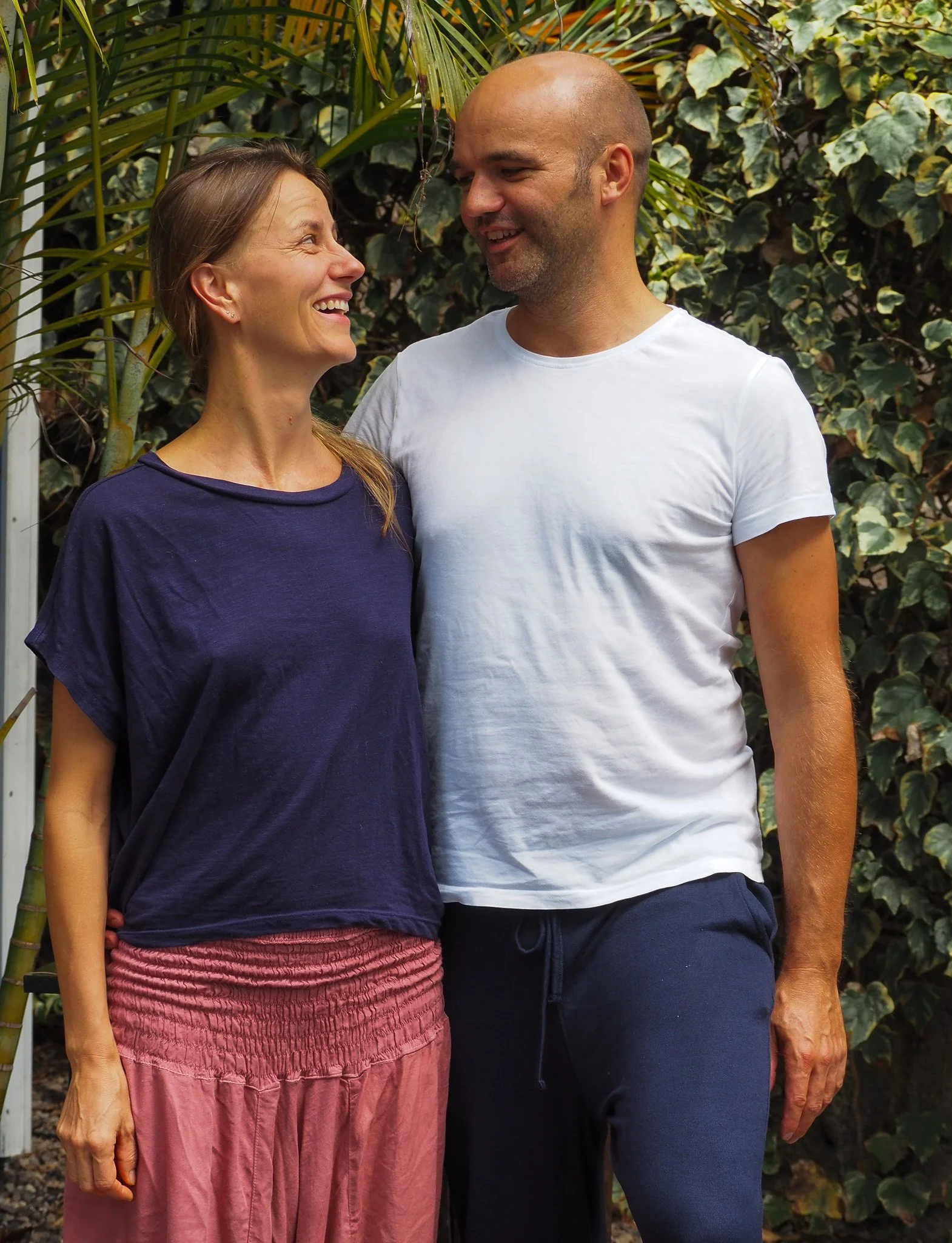 Ein lächelndes Paar steht in einem Garten mit grünen Pflanzen im Hintergrund, sie schauen sich liebevoll an.