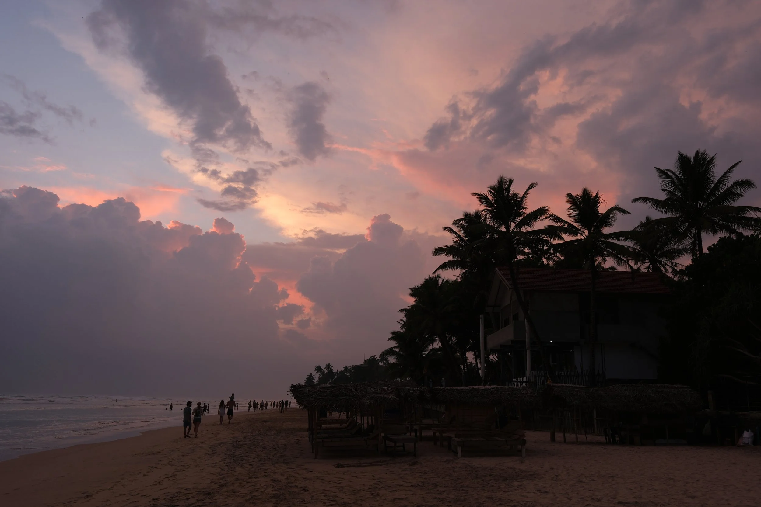 Strand bei Sonnenuntergang mit Palmen, Hütte und Menschen am Wasser