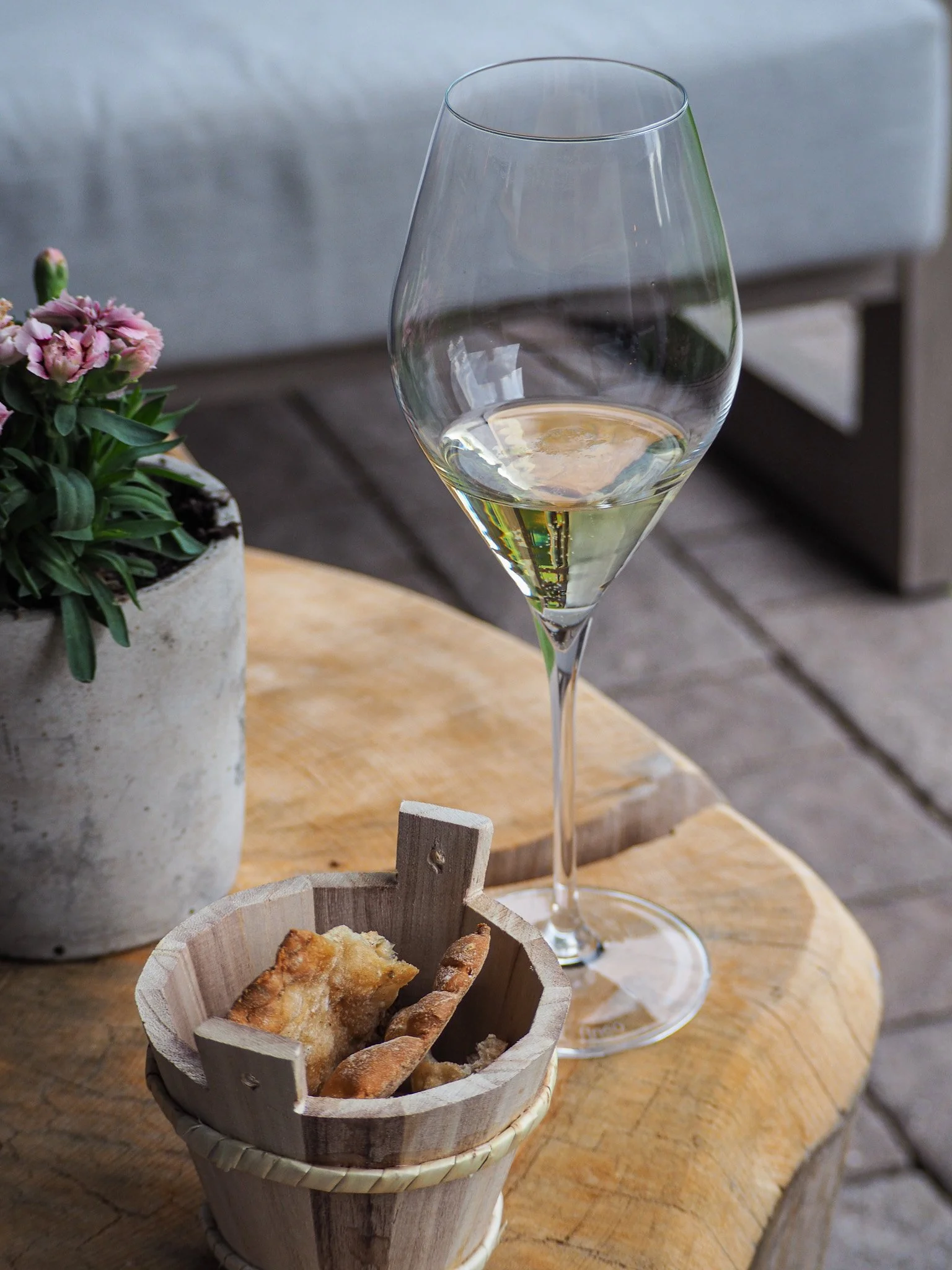 Ein Weinglas mit Weißwein, ein kleiner Korb mit Brot und Fingerfood, eine Topfpflanze mit pinken Blüten auf einem Holztisch, im Hintergrund ein Sofa